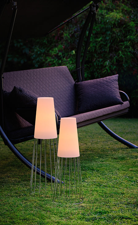 Zwei moderne Stehlampen erzeugen atemberaubende Lichteffekte neben einer gepolsterten Gartenschaukel auf einer Rasenfläche mit üppigem Gartengrün im Hintergrund.