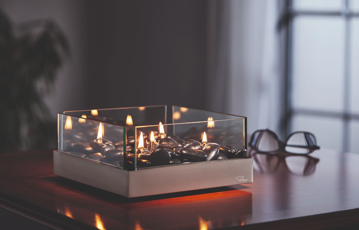 Ein moderner Design-Tischkamin mit Glasseiten und schwarzen Steinen, aus Edelstahl gefertigt und mit Paraffinöl betrieben, steht neben einer Brille auf einem Holztisch.