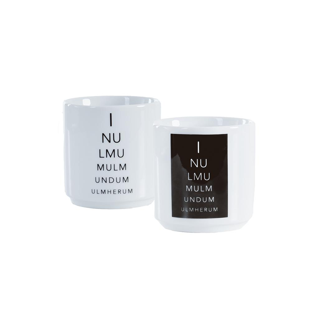 Zwei weiße Ulmer Zungenbrecher-Tassen mit minimalistischem Design stehen nebeneinander und zeigen jeweils Julian Danners Text „I NU LMU MULM UNDUM ULMHERUM“ auf einem schwarzen rechteckigen Etikett, wodurch ein auffälliges monochromes Muster entsteht.