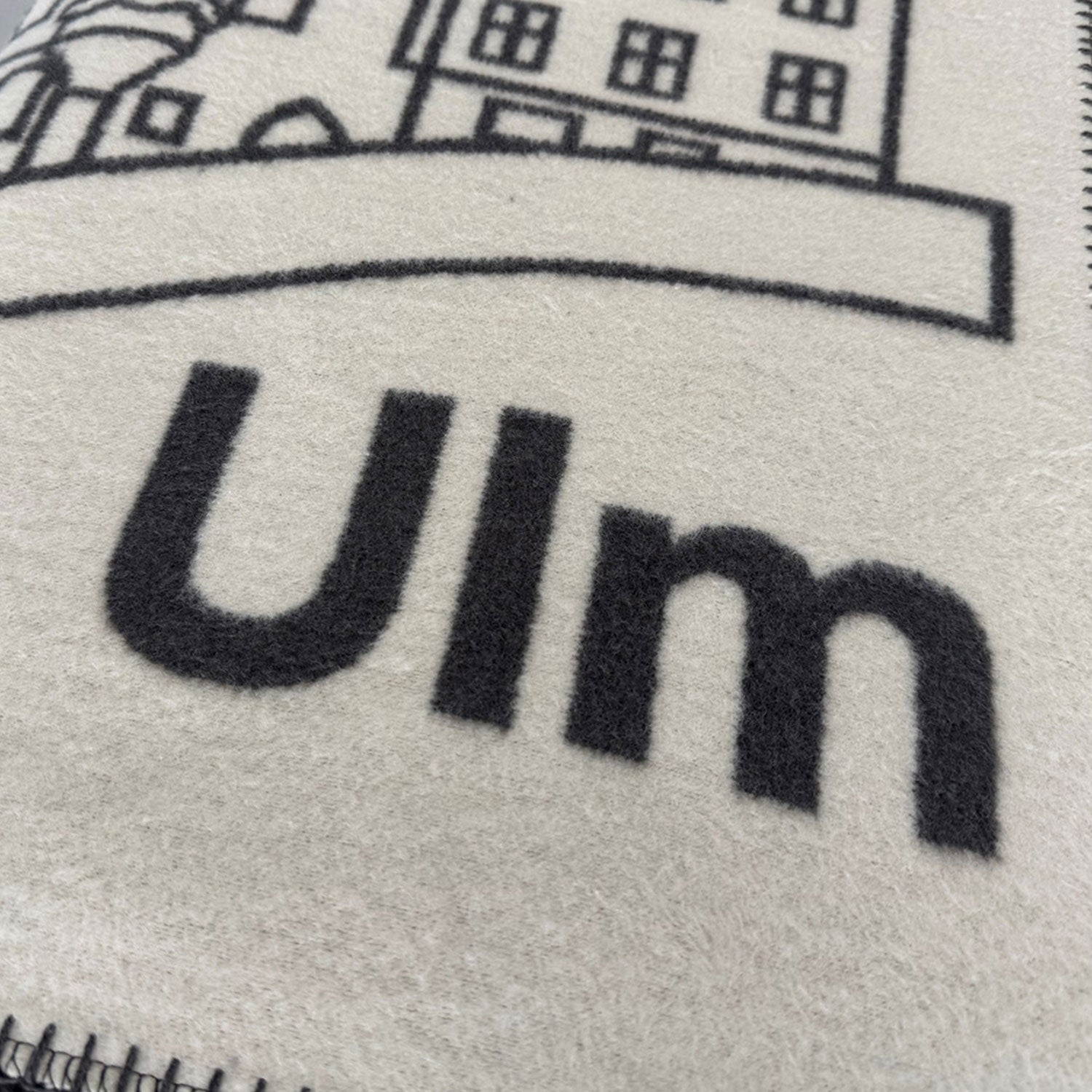 Ein weicher, beigefarbener Stoff zeigt schwarz gestickte Umrisse von Gebäuden, darunter das berühmte Ulmer Münster. Der Schriftzug „Ulm“ ist deutlich unten zu sehen und fängt den Charme dieser Kuscheldecke Ulm aus Baumwolle mit ihrer herrlich gemütlichen und einladenden Textur ein.