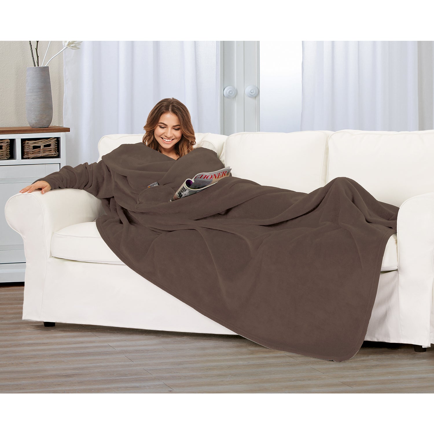 Eine Person entspannt sich auf einem weißen Sofa, eingehüllt in eine Kuscheldecke mit Ärmeln, hält eine Zeitschrift in der Hand und lächelt. Der Raum hat helle Wände, Holzböden und im Hintergrund einen Korb auf dem Regal.