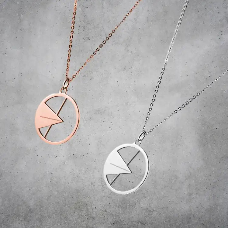 Zwei Schmuckstücke: Die „Ulmer Spatz“ Anhänger und Halsketten mit ovalen Anhängern in Roségold und Silber, jeweils mit geometrischem Blitzmotiv im Oval – präsentiert auf grauem, strukturiertem Hintergrund.