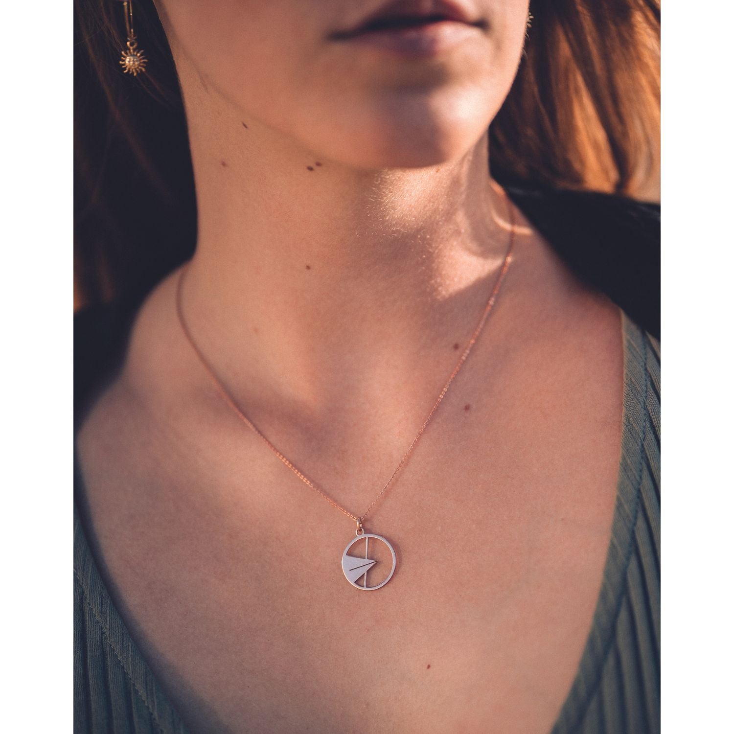 Eine Frau trägt den Anhänger und die Halskette „Ulmer Spatz“ mit rundem Rahmen und Pfeilspitzenform, kombiniert mit einem grünen Faltentop und sonnenförmigen Ohrringen; ihr Gesicht ist teilweise aus dem Rahmen gehoben, wodurch das einzigartige Schmuckstück hervorgehoben wird.