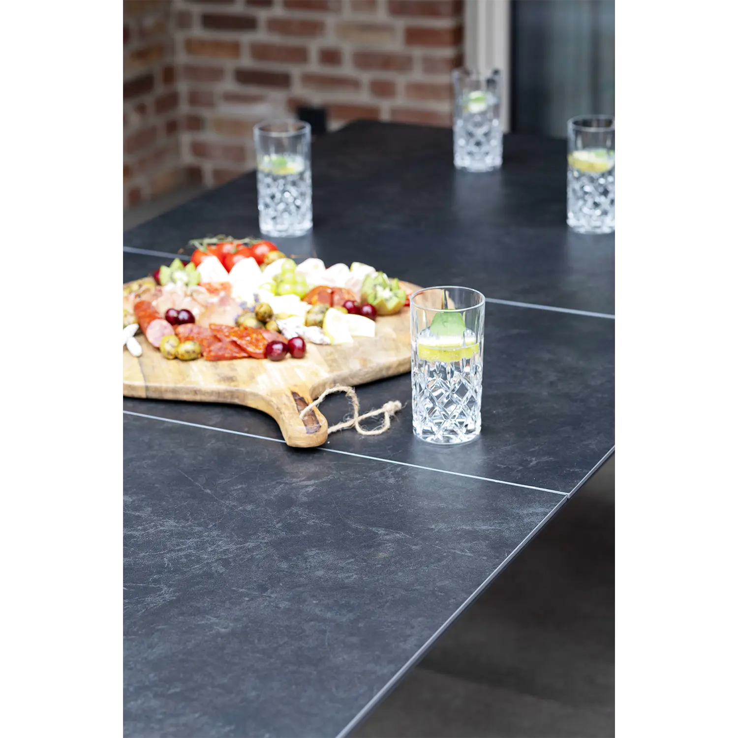Kristallgläser mit Wasser und Limettenscheiben stehen auf dem modernen Ausziehtisch Tarifa. Auf einem Holzbrett werden Käse, Fleisch, Trauben und eingelegtes Gemüse präsentiert. Im Hintergrund ist eine unscharfe Ziegelwand zu sehen.