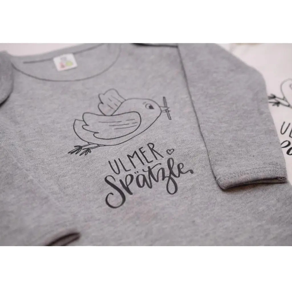 Der Body „Ulmer Spätzle“ ist ein grauer Babybody aus Bio-Baumwolle mit einer minimalistischen Vogel-und-Zweig-Strichillustration und verspieltem „Ulmer Spätzle“-Handlettering unter dem Design.