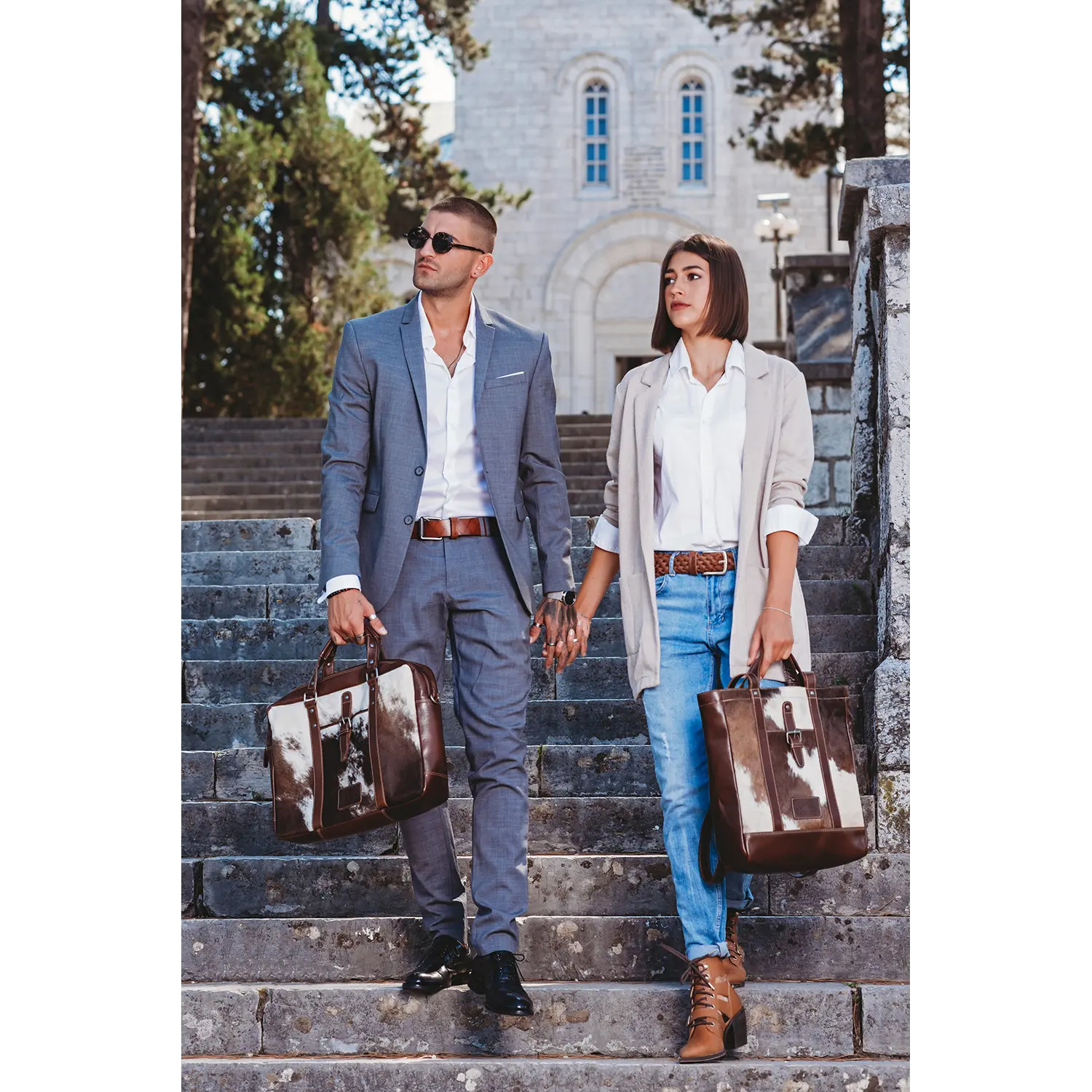Ein stilvolles Paar geht Händchen haltend eine Steintreppe hinunter, beide mit passenden Aktentaschen aus Alpenleder LACKL. Der Mann trägt einen grauen Anzug und eine Sonnenbrille, die Frau einen beigen Blazer und Jeans. Bäume und ein Steingebäude rahmen ihr schickes Ensemble im Hintergrund ein.