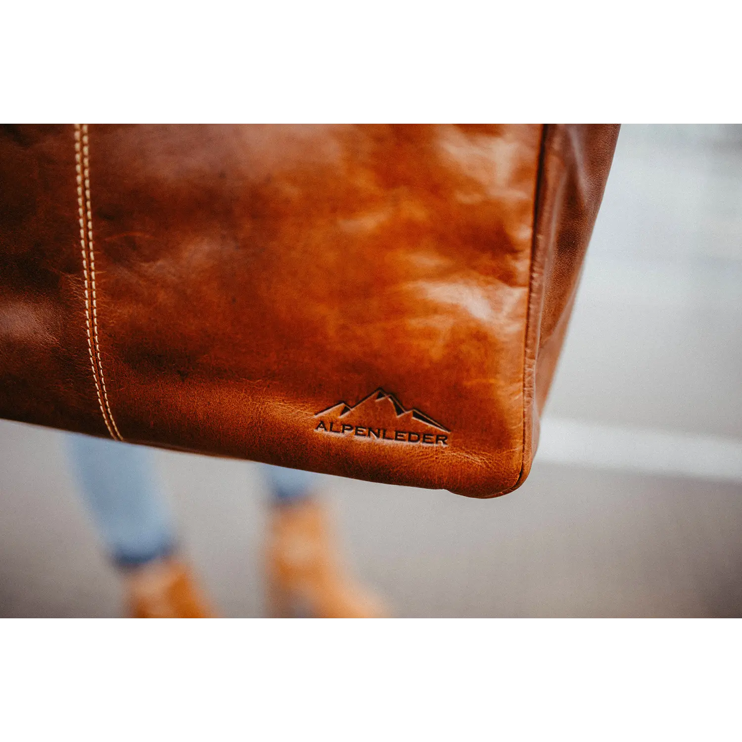 Nahaufnahme der braunen Business-Handtasche ARCO aus Leder mit aufgesticktem Berglogo und „ALPENLEDER“. Der unscharfe Hintergrund zeigt einen Teil eines menschlichen Beins und braunes Schuhwerk und unterstreicht so das sportliche Design der Tasche.