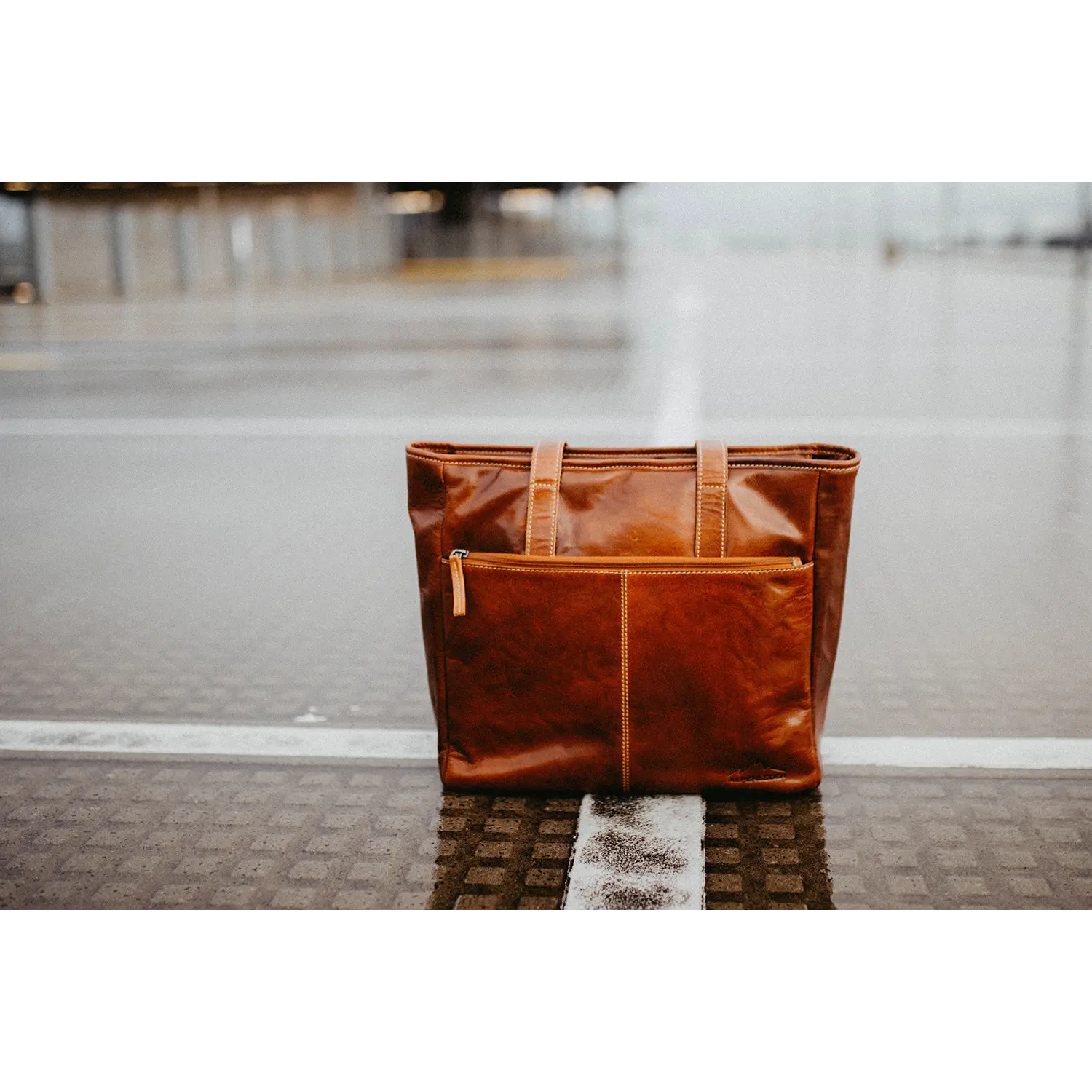 Eine braune Business-Handtasche ARCO aus Leder mit Tragegriffen und Reißverschlussfach vorne steht auf einer nassen, strukturierten Außenfläche. Ihre wasserabweisende Eigenschaft und das Reißverschluss-Hauptfach schützen wichtige Dinge elegant an einem regnerischen Tag, wie der unscharfe Regenguss im Hintergrund suggeriert.