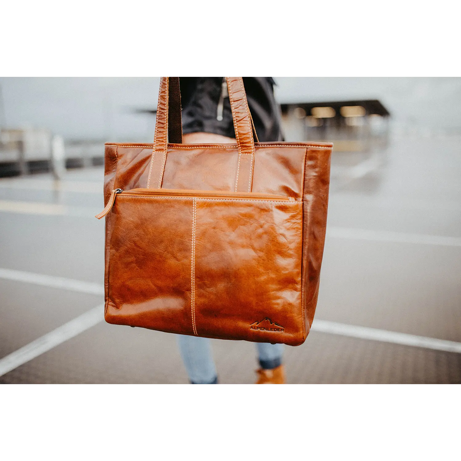 Eine Person hält die Business Handtasche ARCO, eine große braune Ledertasche mit zwei Griffen und einer Reißverschlusstasche vorne. Sie besticht durch sportliche Designelemente, markante Nähte, wasserabweisende Eigenschaften und ein kleines Logo am unteren Rand. Im Hintergrund ist eine verschwommene Szene auf einem Bürgersteig zu sehen.