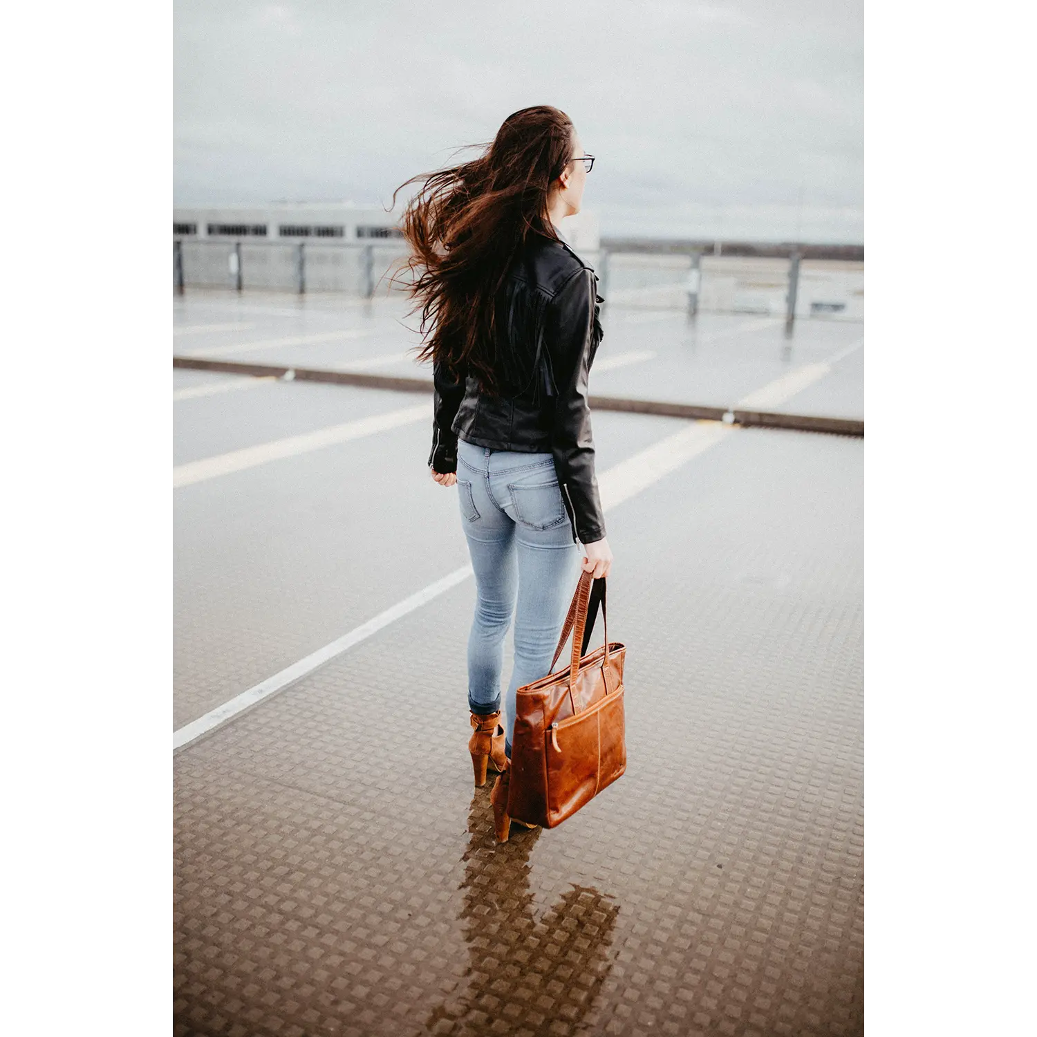 Eine Frau mit langen Haaren steht mit dem Rücken zu einem nassen Dach. Sie trägt eine schwarze Lederjacke und Jeans. Sie trägt die stilvolle braune Business-Handtasche ARCO. Der bewölkte Himmel und der ferne Horizont bilden eine dramatische Kulisse, während das Reißverschluss-Hauptfach ihrer Tasche wichtige Dinge sicher verstaut.