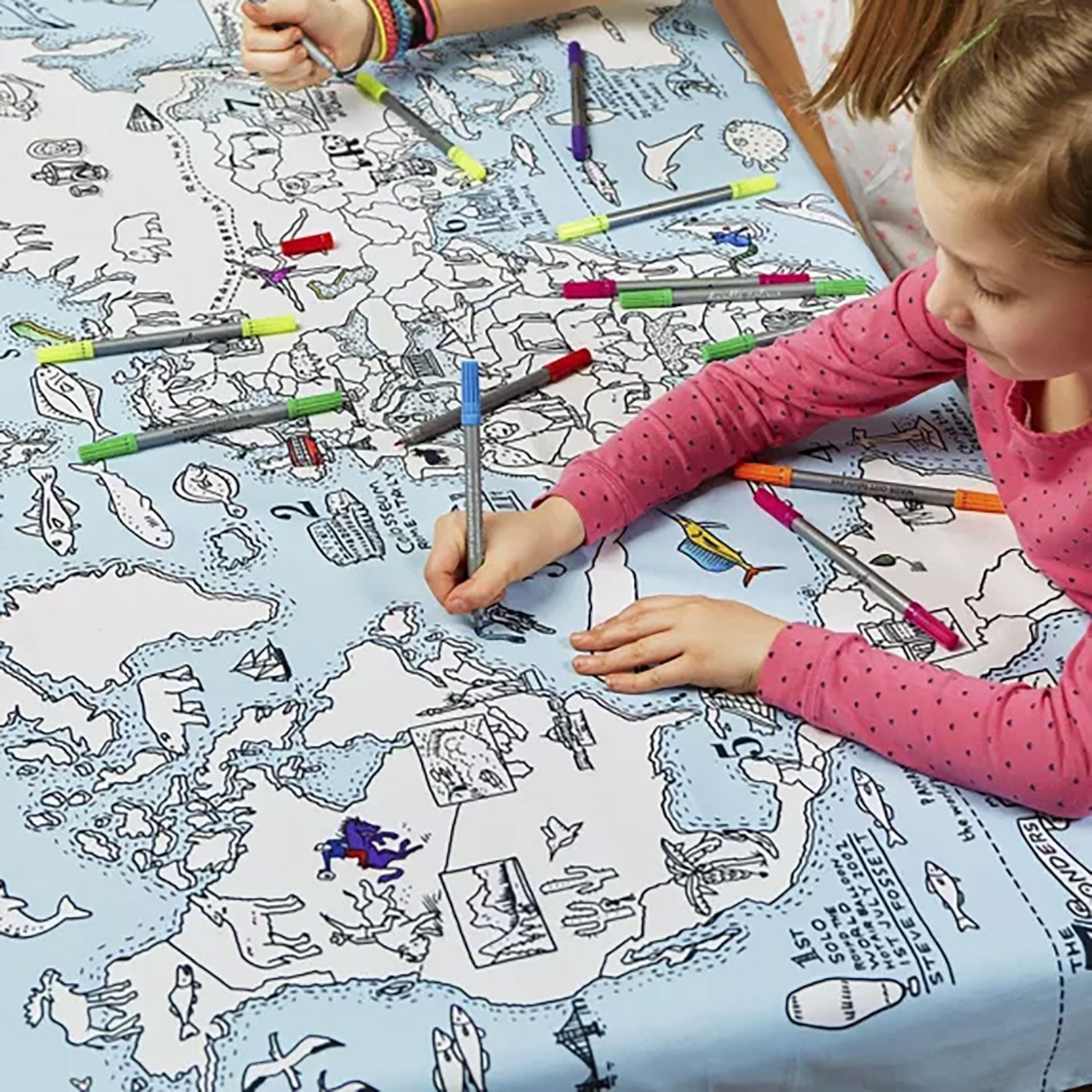 Zwei Kinder bemalen fröhlich eine Baumwolltischdecke mit farbenfrohen geografischen Abbildungen. Ein Kind trägt ein rosa Langarmshirt, und mehrere auswaschbare Stoffmarker liegen verstreut auf dem Tisch.