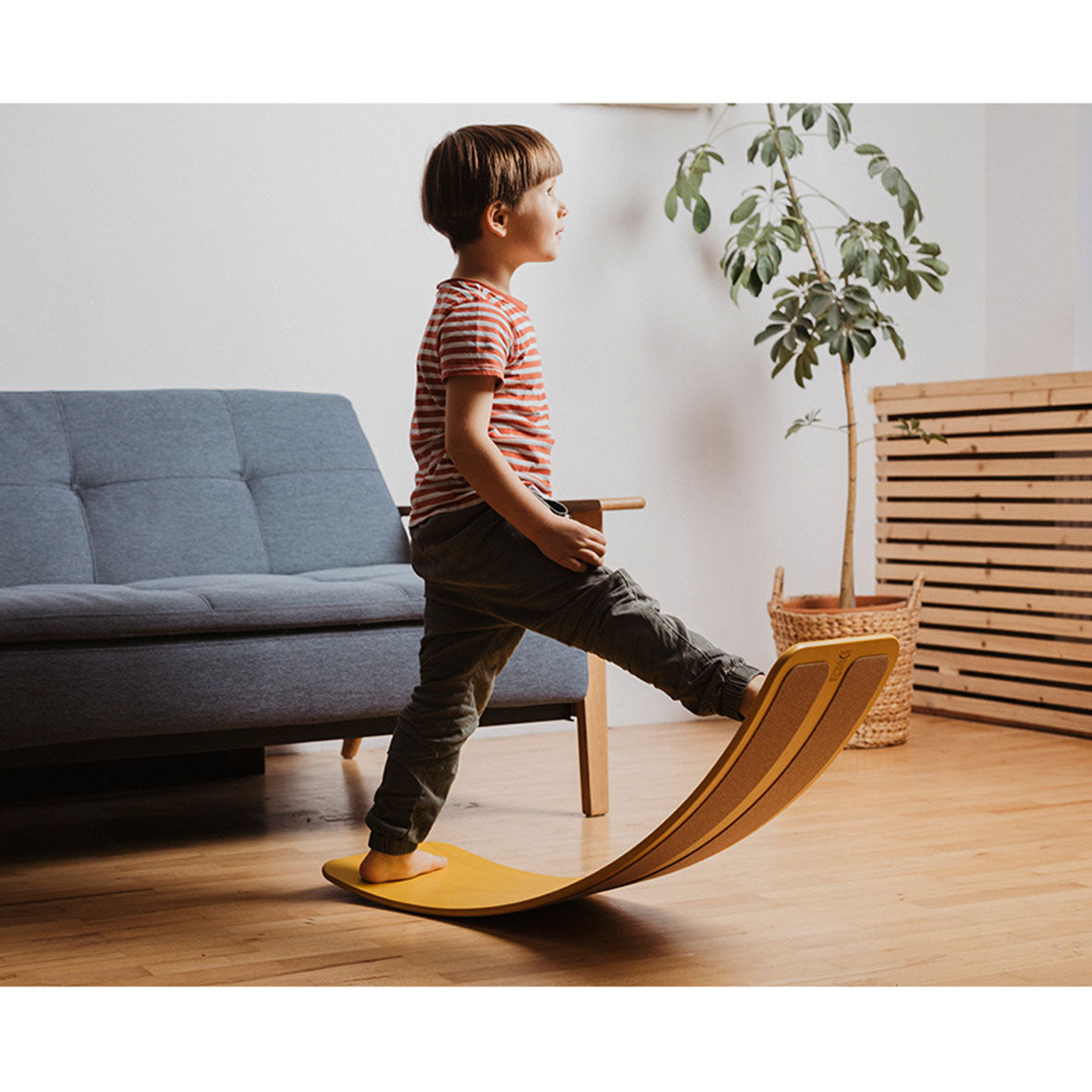 Ein kleines Kind balanciert auf dem das.Brett – Balance Board im Wohnzimmer, ein Fuß angehoben. Im Hintergrund sind ein blaues Sofa, eine grüne Pflanze und Holzmöbel zu sehen. das.Brett ist ein multifunktionales und ökologisches Balance Board.