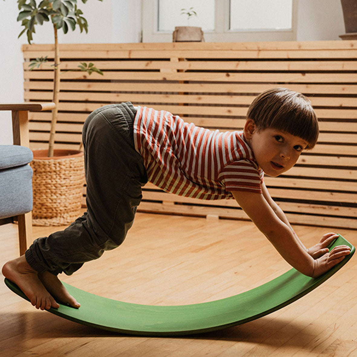 Ein kleines Kind balanciert auf einem grünen Balance Board im Innenbereich. Hände und Füße stehen auf dem Brett, die Hüften sind angehoben. Das Kind trägt ein rot gestreiftes Hemd und eine graue Hose. Holzmöbel und eine Topfpflanze bereichern den multifunktionalen Raum.