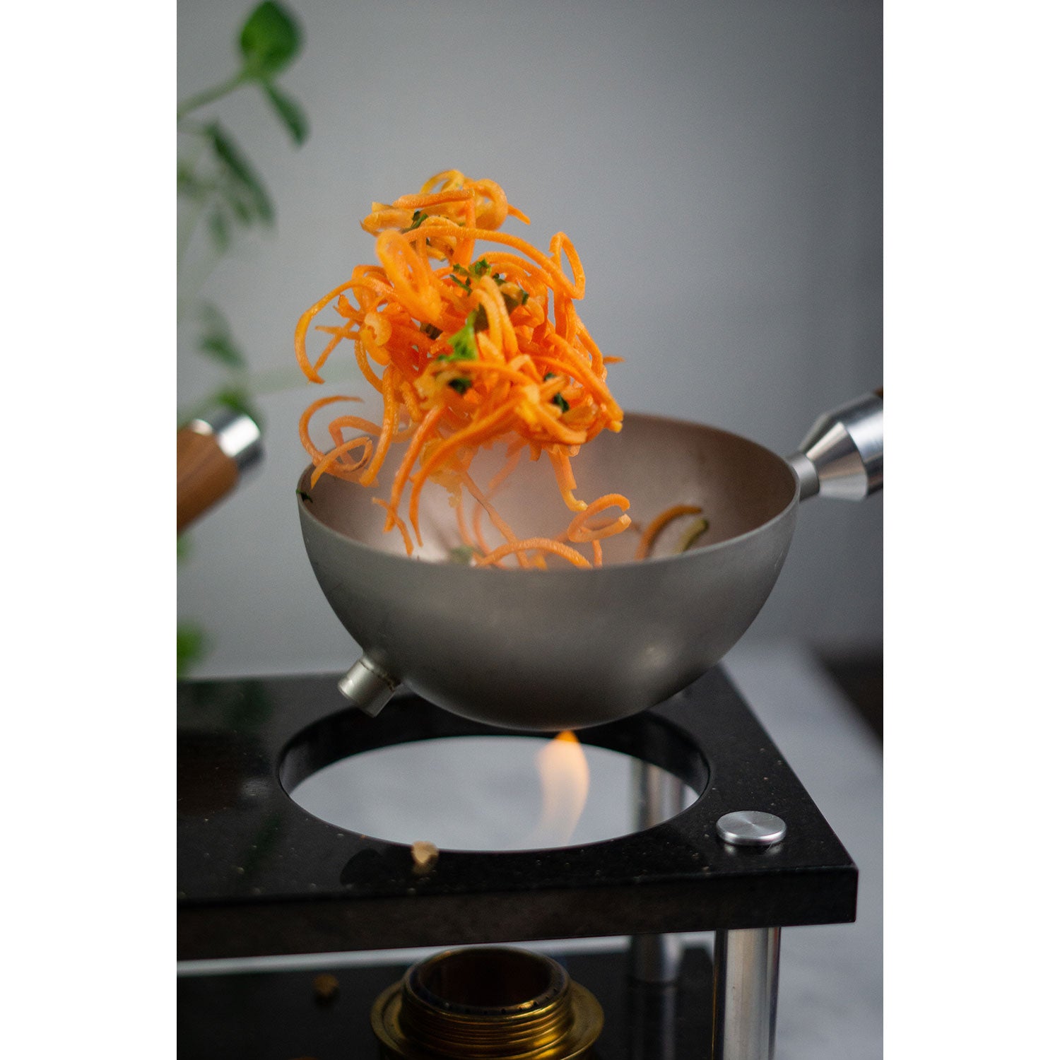Eine dynamische Aufnahme zeigt leuchtend orangefarbenes, spiralförmiges Gemüse, das in einem grauen, halbkugelförmigen DeWok in der Luft herumgeworfen wird. Das neue Kochsystem! Über einer kleinen Flamme auf einer schwarzen Arbeitsplatte positioniert – für den perfekten Soulfood-Moment, mit dezent grünem Laub im Hintergrund.