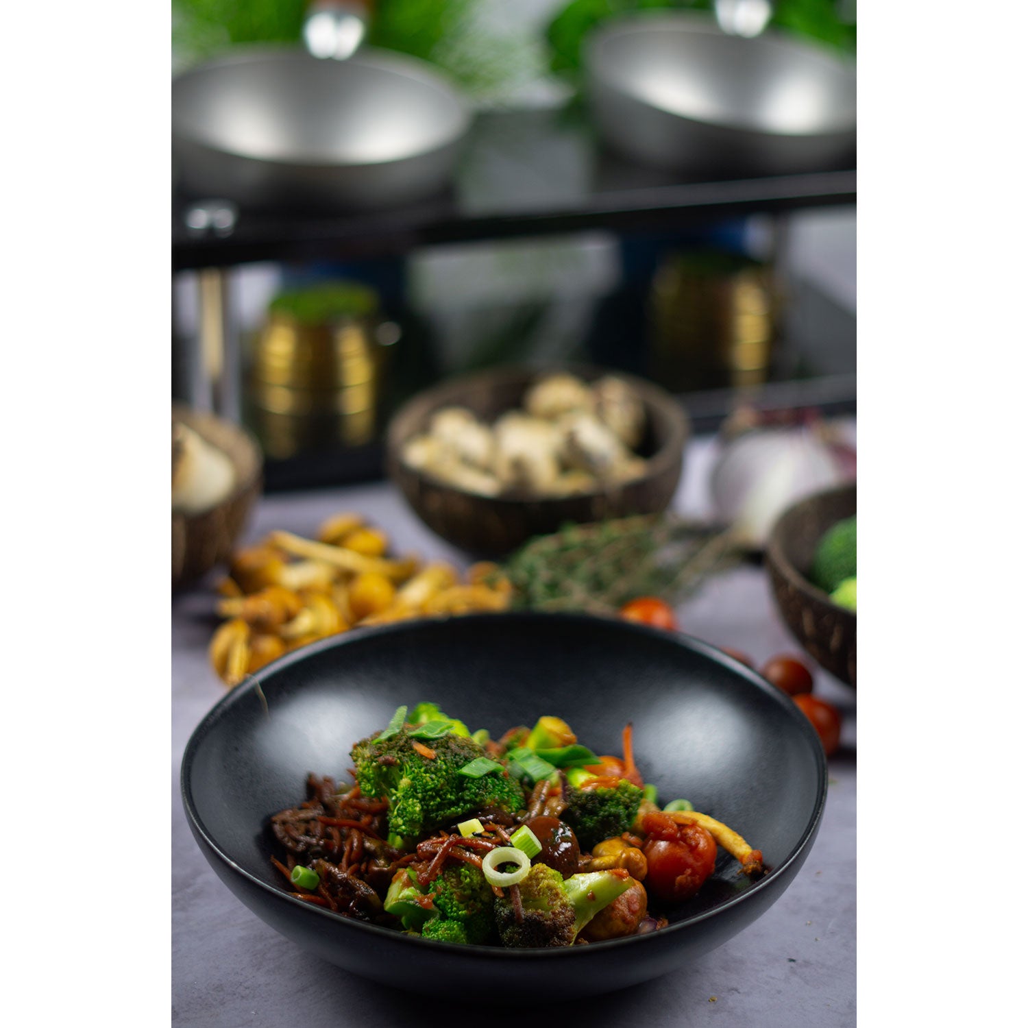 Eine schwarze Schüssel mit gebratenem Gemüse, Rindfleisch und Frühlingszwiebeln, garniert mit Sesam, fängt einen Soulfood-Moment ein. Verschwommene Schalen mit Pilzen und Tomaten im Hintergrund begleiten dieses genussvolle DeWok-Erlebnis mit „DeWok. Das neue Kochsystem!“.