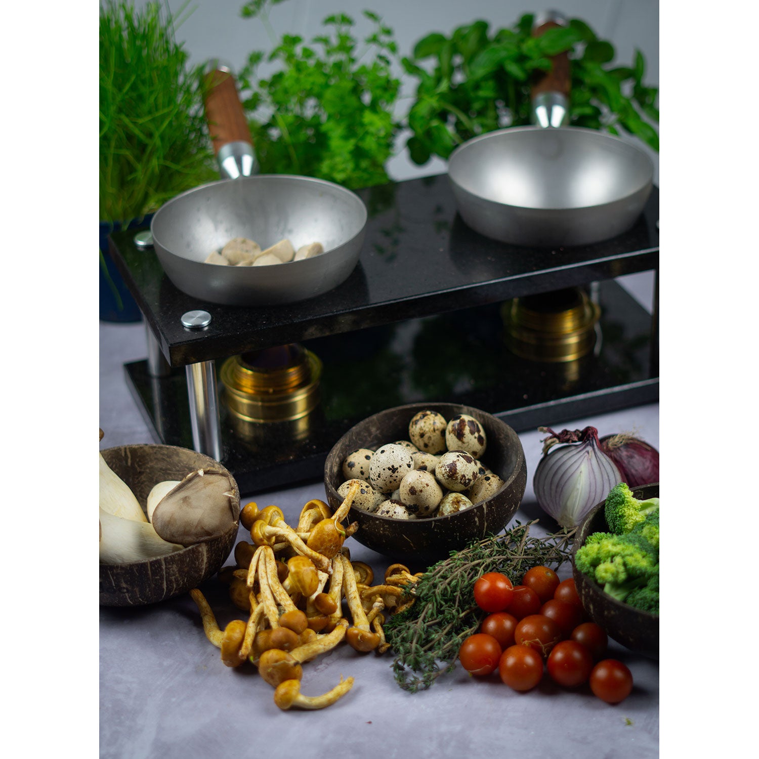 Ein DeWok-Kochsetup mit zwei kleinen Pfannen auf schwarzem Kochfeld, umgeben von frischen Kräutern, Pilzen, Wachteleiern, Kirschtomaten, Brokkoli, Zwiebeln und Thymian auf grauer Fläche. Ideal für einen Soulfood-Moment.
