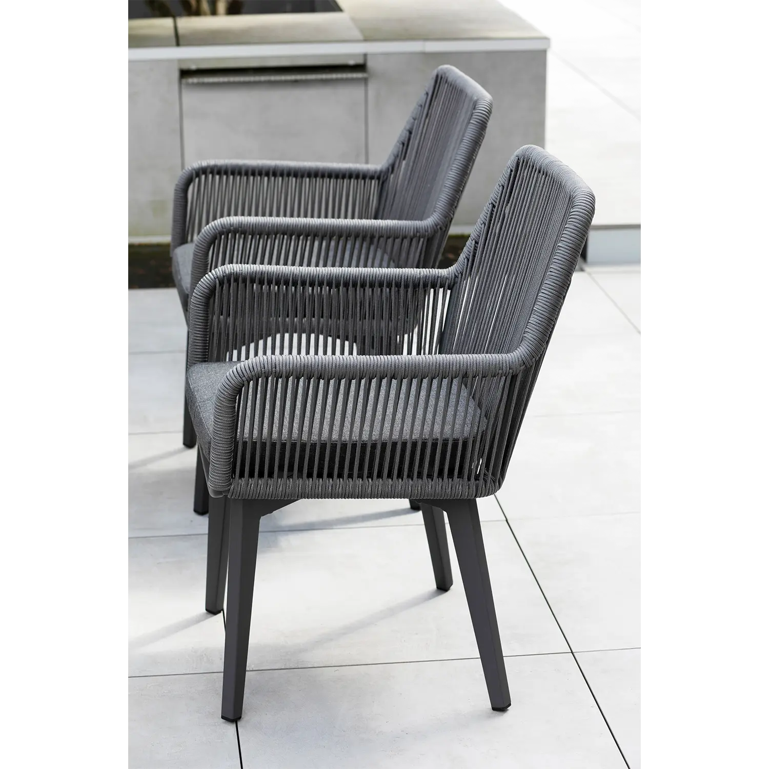 Das Esszimmersessel Marbella Rope 2er Set mit dunkelgrauem Seilgeflecht und Holzbeinen steht elegant auf einer hellgrau gefliesten Terrasse. Sein minimalistisches Design harmoniert mit der Betonumgebung und bietet eine stilvolle und wetterfeste Wahl für den Außenbereich.