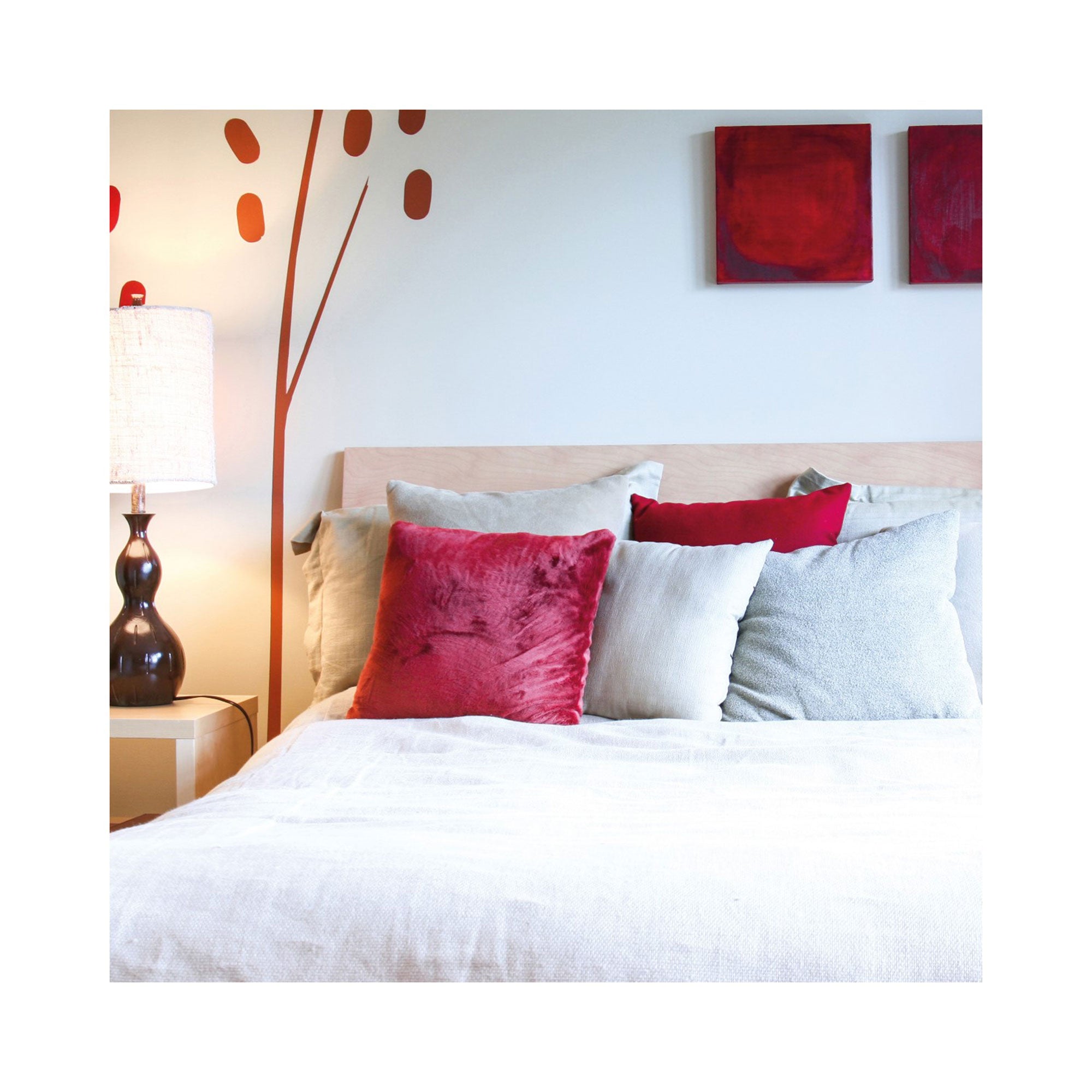 Ein modernes Schlafzimmer mit Holzbett, weißer Bettwäsche und rotgrauen Kissen vermittelt die perfekte Atmosphäre der „Erlebnis-Box 3 Tage Du & Ich“. Ein Nachttisch aus Holz trägt eine dunkle Lampe, während zwei minimalistische rote Kunstwerke die Wand daneben schmücken.