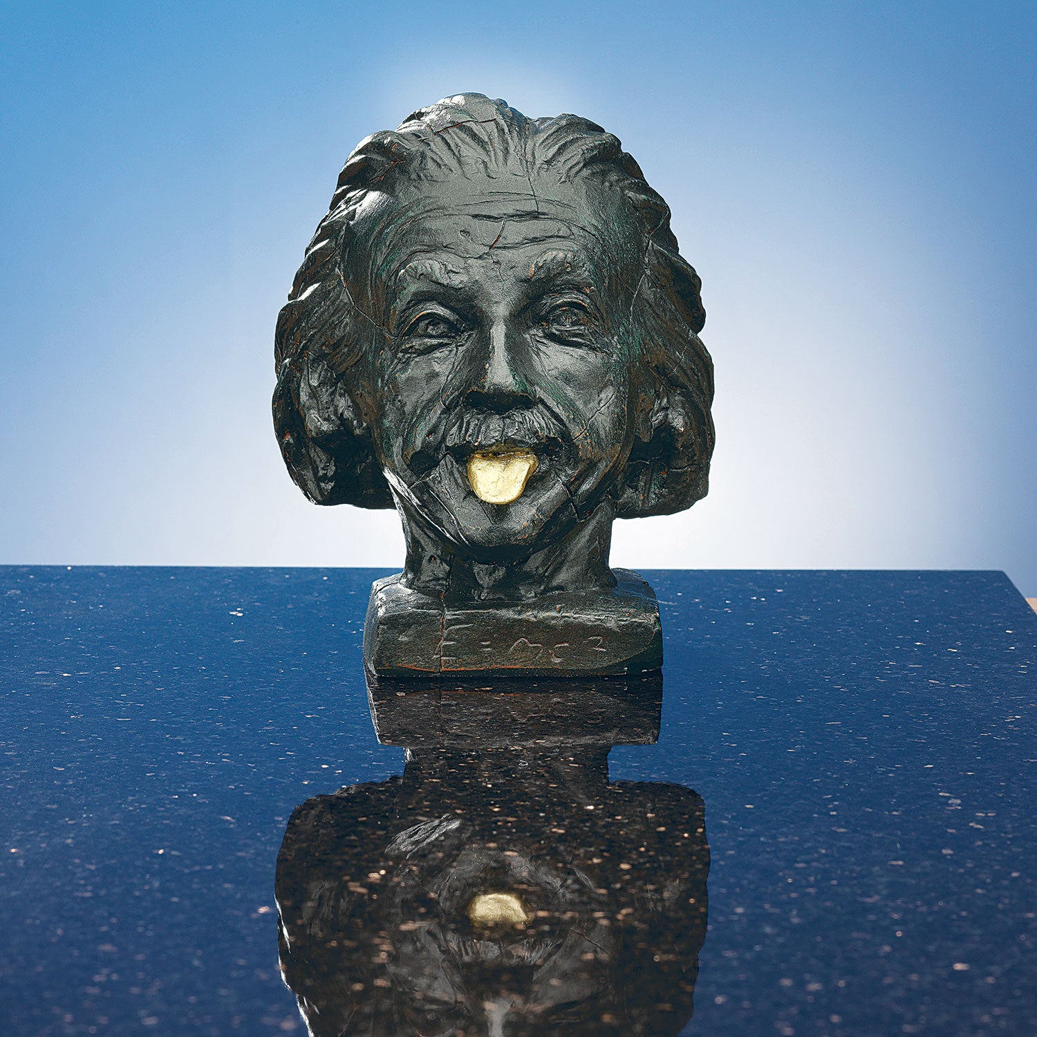 Die Skulptur „Einstein-Kopf mit goldener Zunge“ zeigt den berühmten Wissenschaftler mit herausgestreckter Zunge auf einer glänzenden Oberfläche vor einem hellblauen Hintergrund mit Farbverlauf.