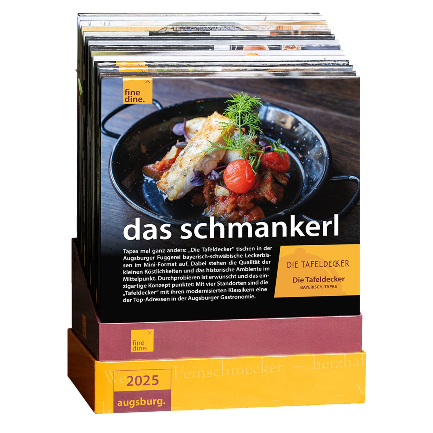 Ein Schauständer präsentiert deutsche Restaurantzeitschriften mit dem Titel „Das Schmankerl“, deren Cover gegrillten Fisch, Kirschtomaten und Gemüse zeigt, und dem Untertitel „Die TafelDecker“. Entdecken Sie kulinarische Erlebnisse mit der Fine Dine Box Augsburg 2025.