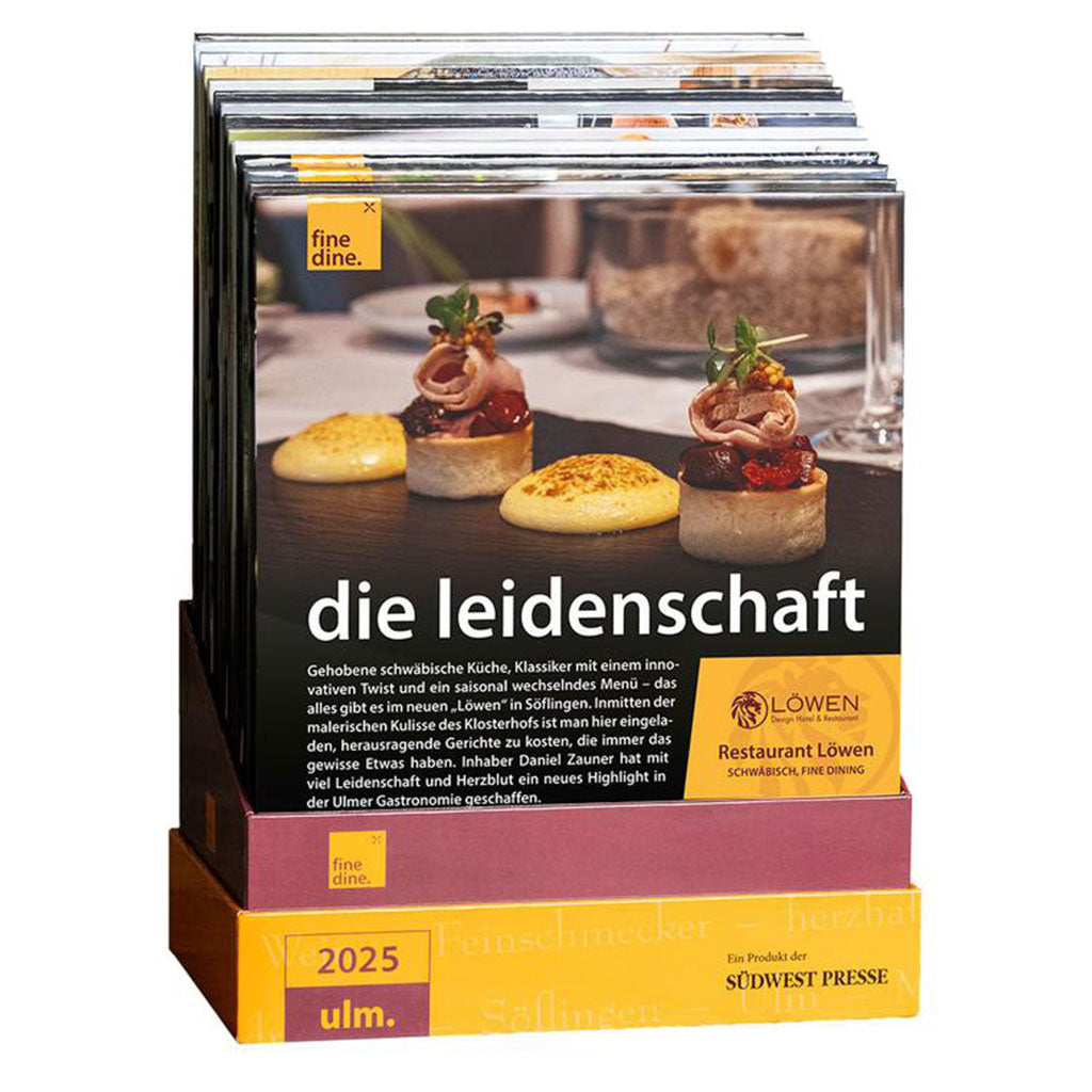 Ein Stapel kulinarischer Zeitschriften der „Fine Dine Box Ulm 2025“ mit Gourmet-Gerichten und Einblicken aus dem Restaurant Löwen liegt übersichtlich auf einem lila-gelben Ständer. Perfekt für alle, die kulinarische Erlebnisse in Ulm suchen.