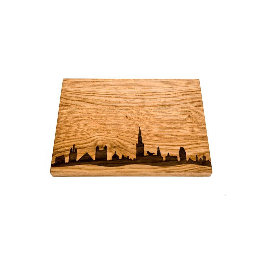 Das handgefertigte „Frühstück- oder Vesperbrettchen – hell“ zeigt ein geätztes Skyline-Design mit verschiedenen Gebäuden und einer markanten Turmspitze aus hellem Eichenholz, das eine schöne natürliche Holzmaserung vor einem weißen Hintergrund hervorhebt.
