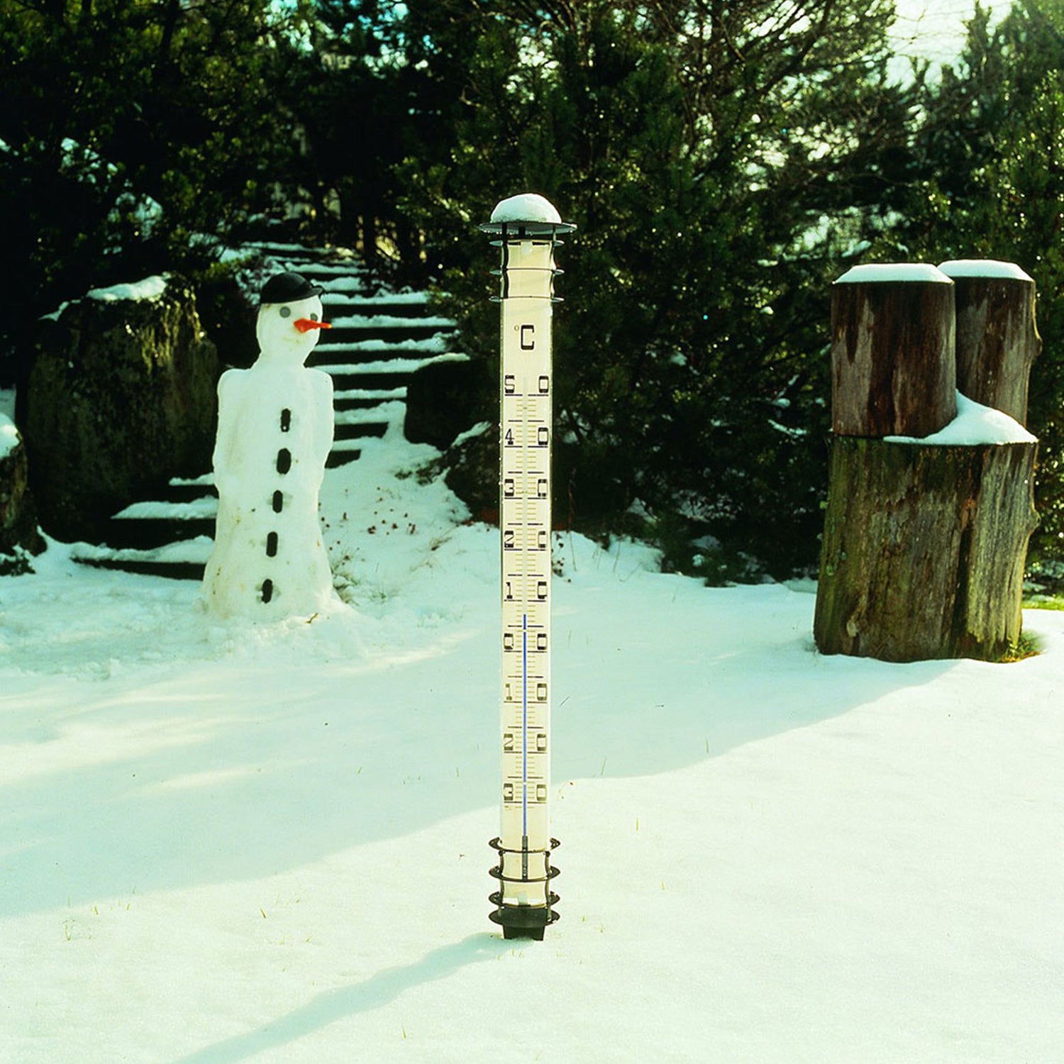 In einem schneebedeckten Garten sind ein Schneemann und eine Holzkonstruktion mit Bäumen unter klarem Himmel zu sehen. Im Vordergrund misst ein hohes Gartenthermometer auf einem Erdspieß die Schneehöhe.