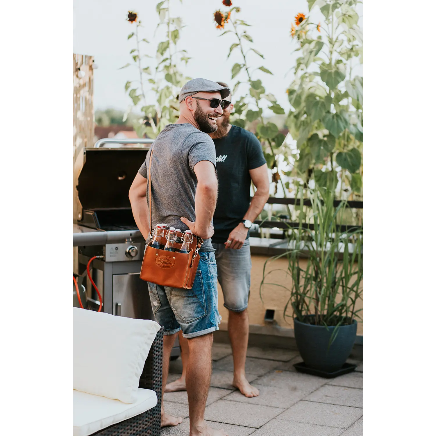 Zwei Männer stehen auf einer Terrasse mit Sonnenblumen im Hintergrund. Einer trägt eine graue Kappe und eine Sonnenbrille und hält stolz seine mit Flaschen gefüllte Getränketasche ALPENSECHSER-Harness in der Hand. Sie stehen in der Nähe eines Grills und Terrassenmöbeln und genießen den sonnigen Tag.