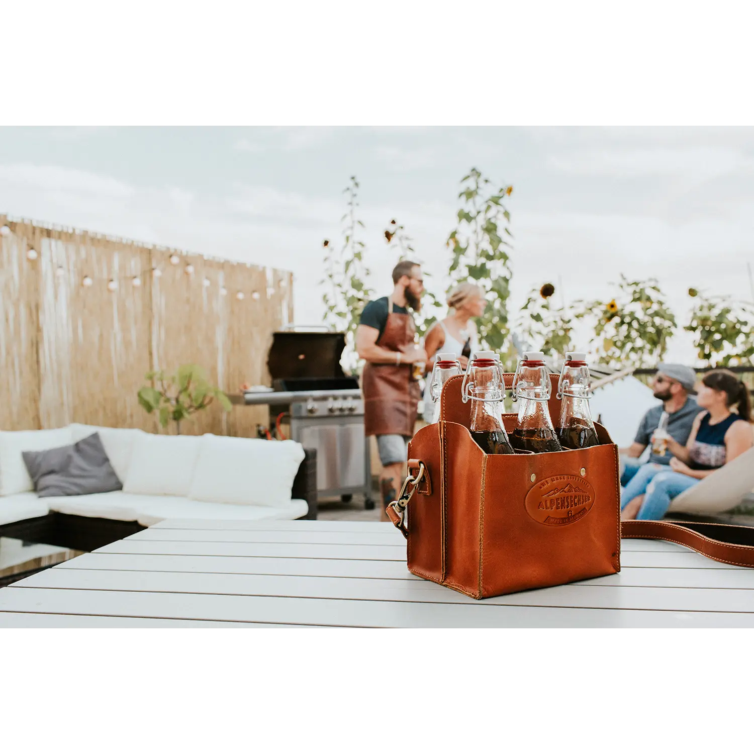 Eine mit Flaschen gefüllte Getränketasche ALPENSECHSER-Harness steht auf einem weißen Tisch, während sich drei Personen bei einem Grill auf der Terrasse unterhalten, umgeben von Gartenmöbeln und einem Sichtschutz aus Bambus unter klarem Himmel.