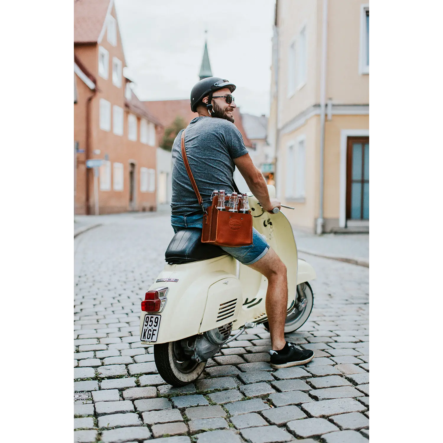 Eine Person mit Helm und Sonnenbrille sitzt auf einem weißen Oldtimer-Roller, der auf einer Kopfsteinpflasterstraße geparkt ist. Über der Schulter hängt eine Getränketasche von ALPENSECHSER. Die Straße, gesäumt von pastellfarbenen Gebäuden, verströmt zeitlosen Charme.