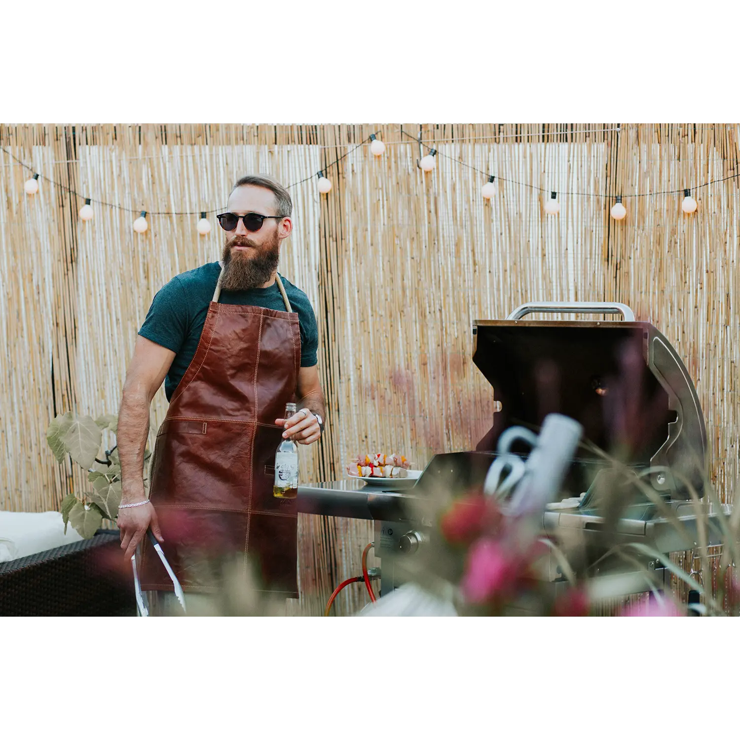 Ein bärtiger Mann mit Sonnenbrille und Alpenleder-Grillschürze FUNKENFEUER steht neben einem Grill und hält ein Getränk und eine Grillzange in der Hand. Lichterketten funkeln darüber vor einem Bambuszaun. Neben ihm steht ein Tisch mit Spießen, der eine einladende Festtagsatmosphäre im Freien schafft.