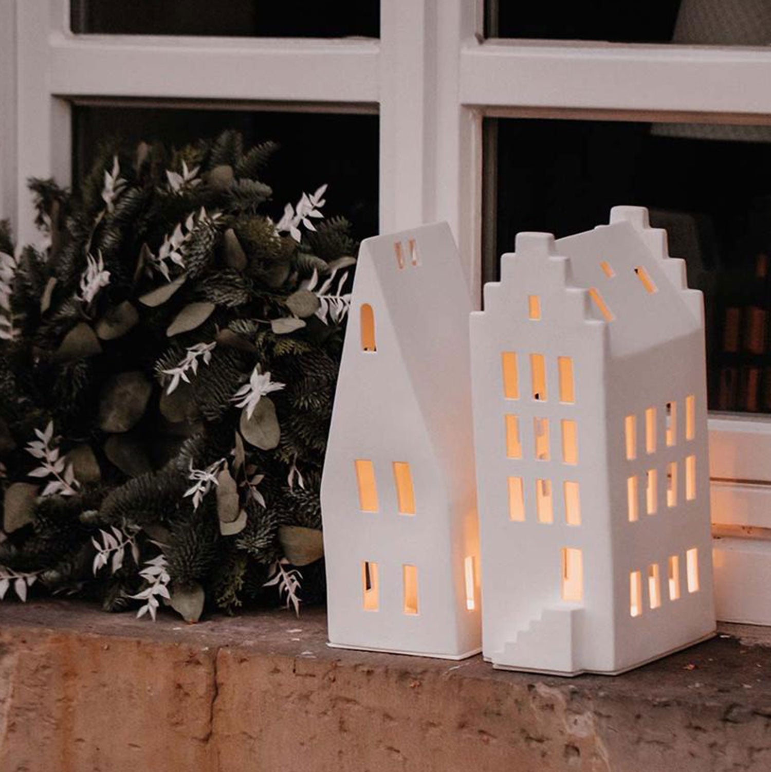 Zwei unglasierte Große Lichthäuser „Giebeldach“ aus Porzellan, aus deren Fenstern warmes Licht strahlt, stehen auf einem Fenstersims. Ein üppiger grüner Kranz steht daneben und verbreitet eine gemütliche Atmosphäre und festliche Stimmung.