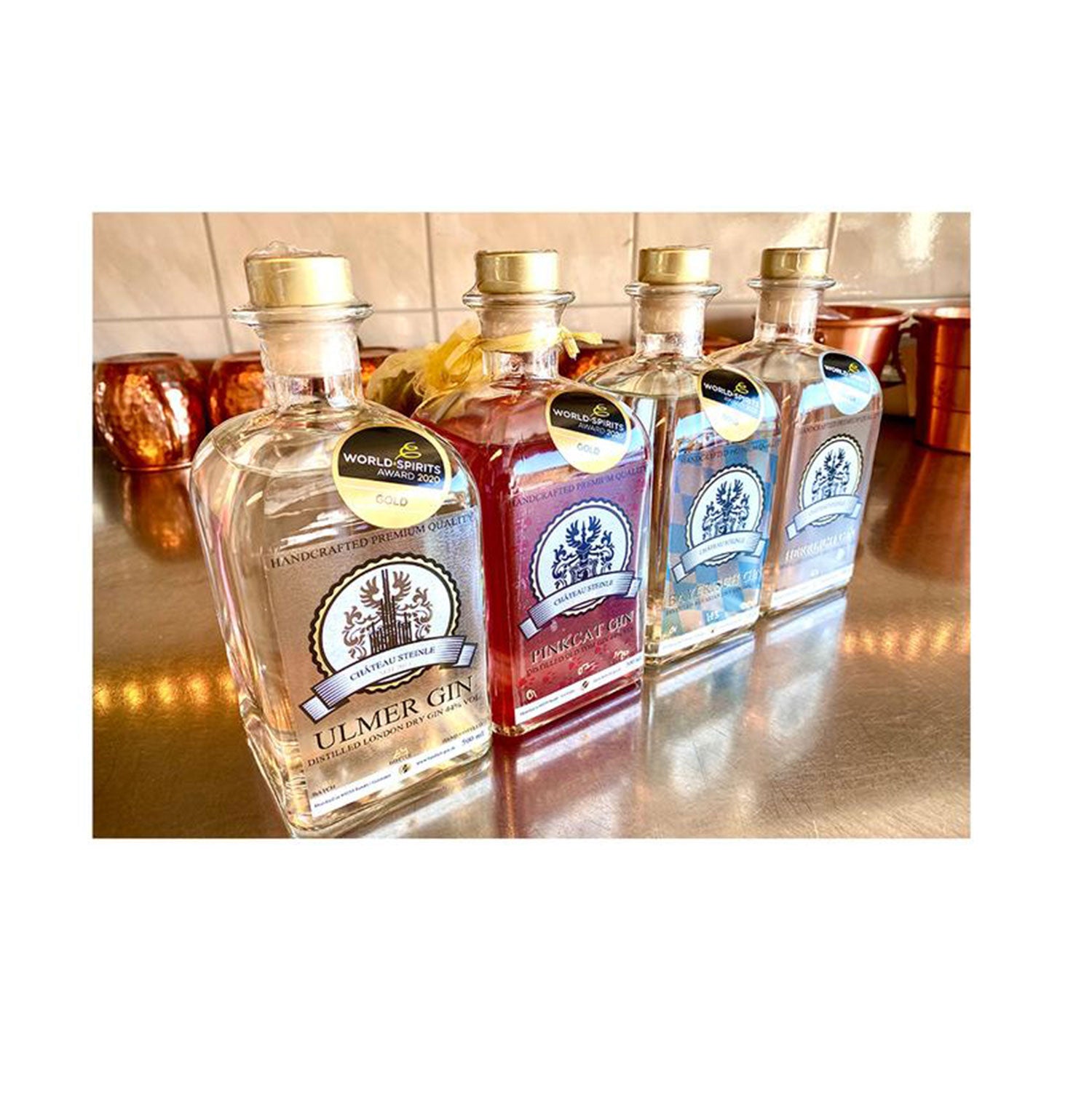 Vier Flaschen des Herrlich Gin Geschenksets mit einzigartigen Etiketten stehen auf einer reflektierenden Oberfläche. Die mittlere Flasche ist rot, wie Pinkcat Gin, während die anderen klar sind. Jede Flasche ist mit Flaschenhalsanhängern und einem Baumlogo versehen, alles vor kupfernen Elementen im Hintergrund.
