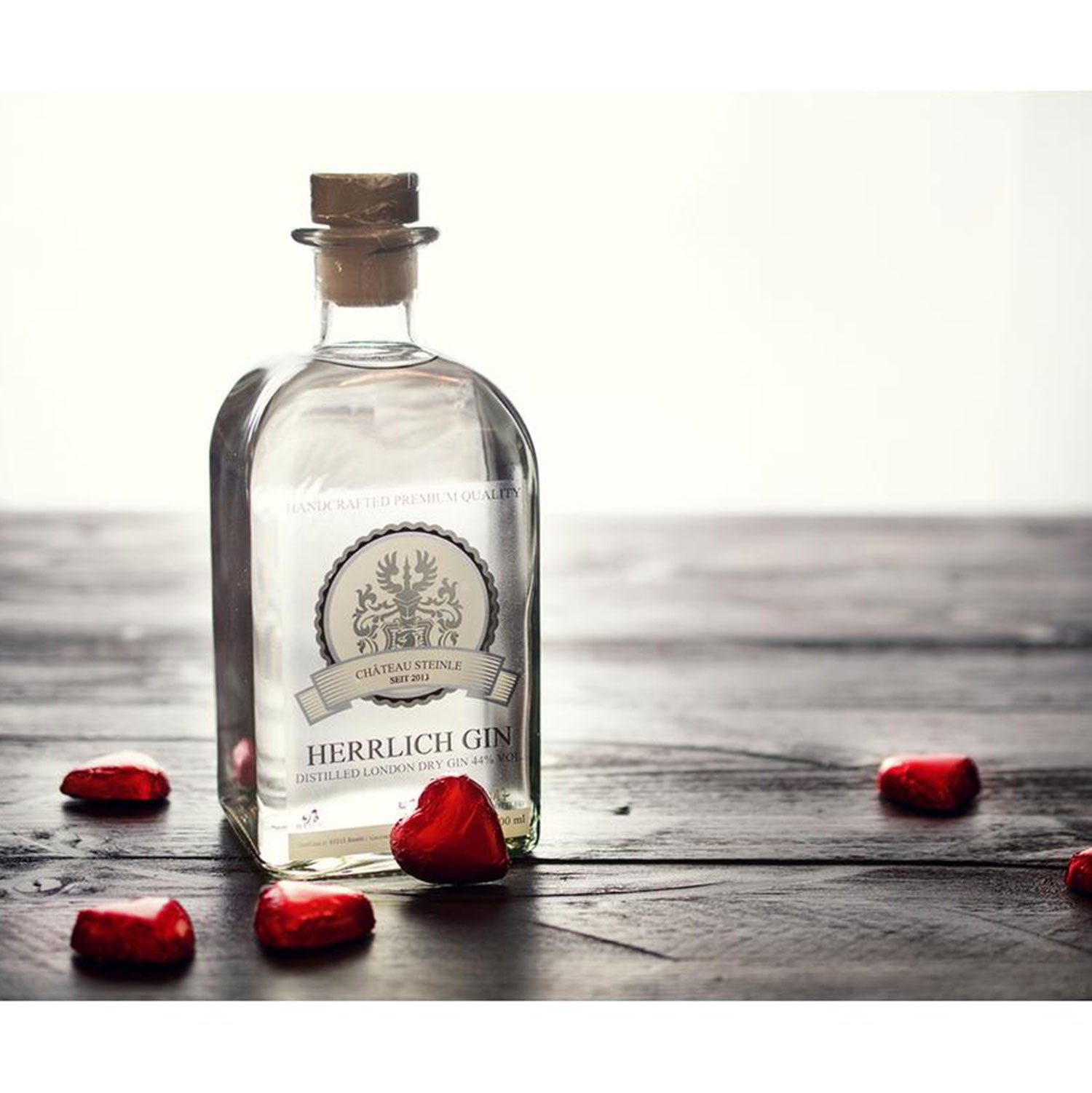 Eine mit Korken verschlossene Herrlich Gin-Flasche, gefertigt im Stil eines London Dry Gin, steht auf einem Holztisch. Rote Schokoladenherzen aus Folie umrahmen sie, während sanfte Beleuchtung und dunkles Holz die regionale Herstellung betonen.