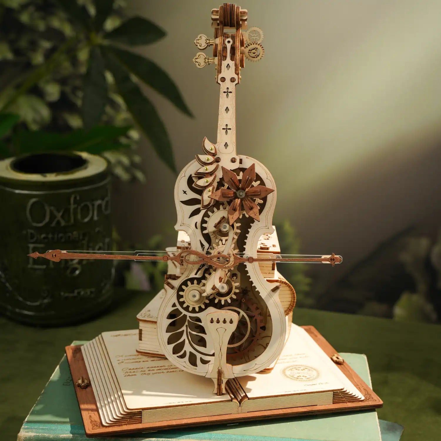 Das 3D Holzpuzzle „Magisches Cello“, ein Holzmodell mit filigranen Zahnrädern und floralen Mustern, steht auf einem Tisch. Dahinter steht ein grüner, zylindrischer Behälter im Vintage-Stil mit der Aufschrift „Oxford“, umgeben von üppigem Laub – eine wahre Freude für jeden Musikliebhaber.