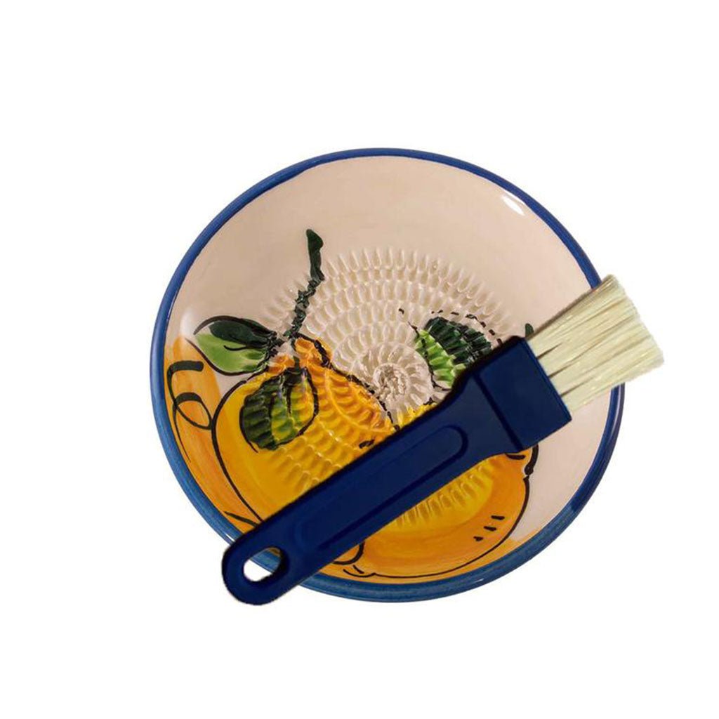 Eine kleine Keramikreibe – gelb mit aufgemaltem Zitronen-Design und erhabener Textur. Ein Backpinsel mit blauem Griff und beigen Borsten befindet sich in diesem Haushaltshelfer. Der weiße Hintergrund hebt die farbenfrohen Details wunderschön hervor.
