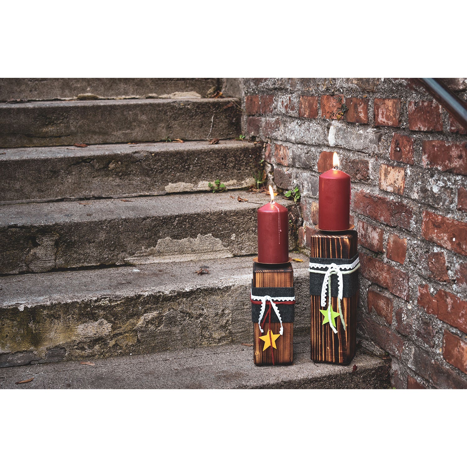 Zwei rote Stumpenkerzen, beleuchtet auf Kerzenständern aus Holz – groß mit Sternendekor, beleuchten die Stufen neben einer roten Backsteinmauer. Der verwitterte Stein trägt zum rustikalen Ambiente bei.