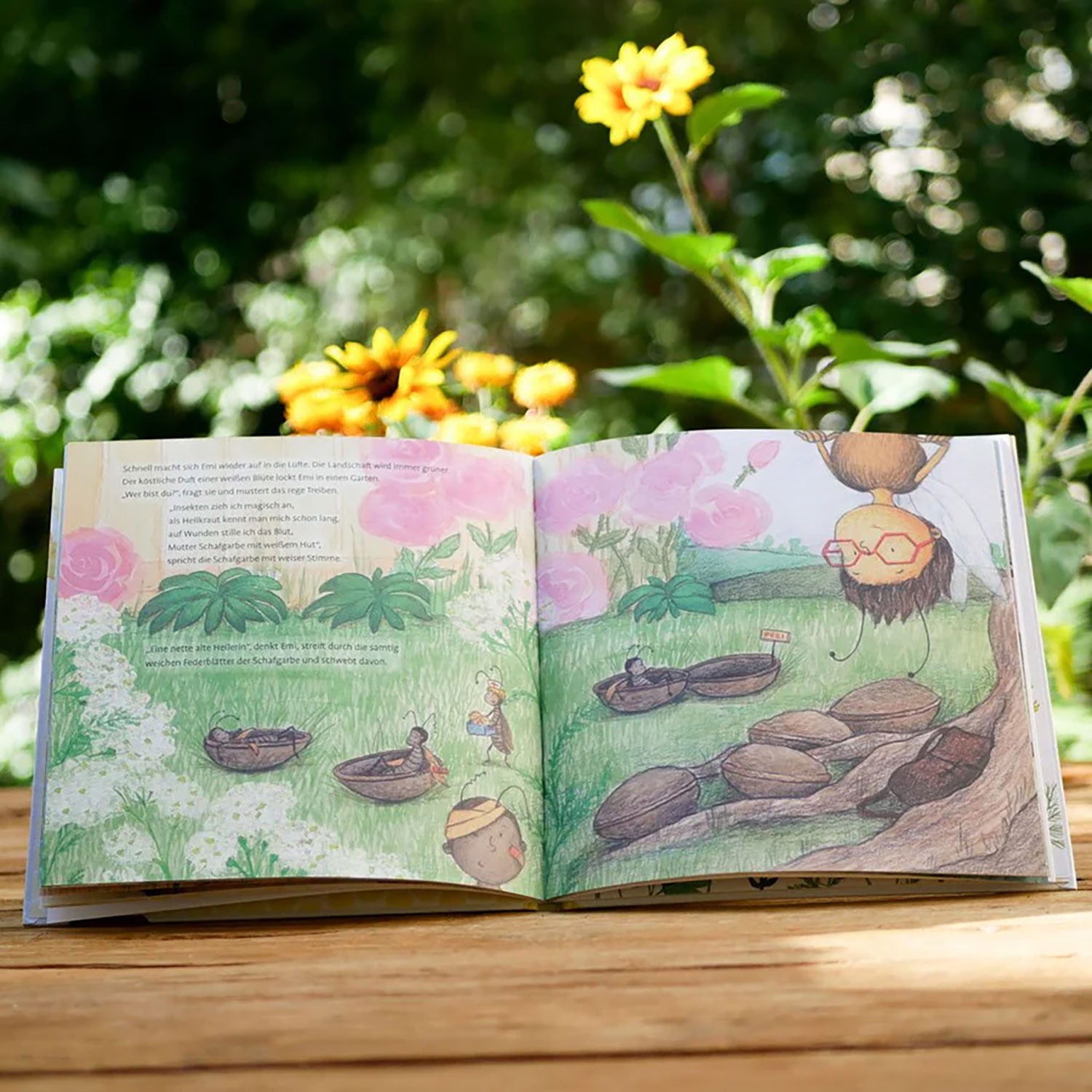 Ein aufgeschlagenes Exemplar von „Emi Brillenbiene – Die weite Reise einer kleinen Biene“ liegt auf einem Holztisch im Freien, umgeben von üppigen grünen Pflanzen und Sonnenblumen. Bunte Illustrationen zeigen Honigbiene Emi beim Erkunden bienenfreundlicher Blumen, gepaart mit skurrilem Text.