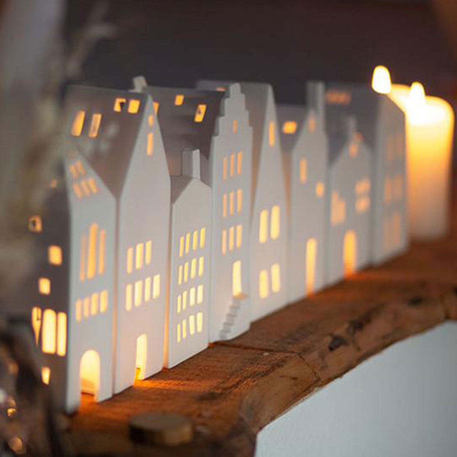 Die Miniaturen „Spitzdach“ von Kleines Lichthaus mit beleuchtetem Innenleben schmücken ein Holzregal, während Kerzen im Hintergrund ein warmes Licht ausstrahlen und so eine gemütliche Atmosphäre schaffen, die ideal für Weihnachtsfeiern ist.