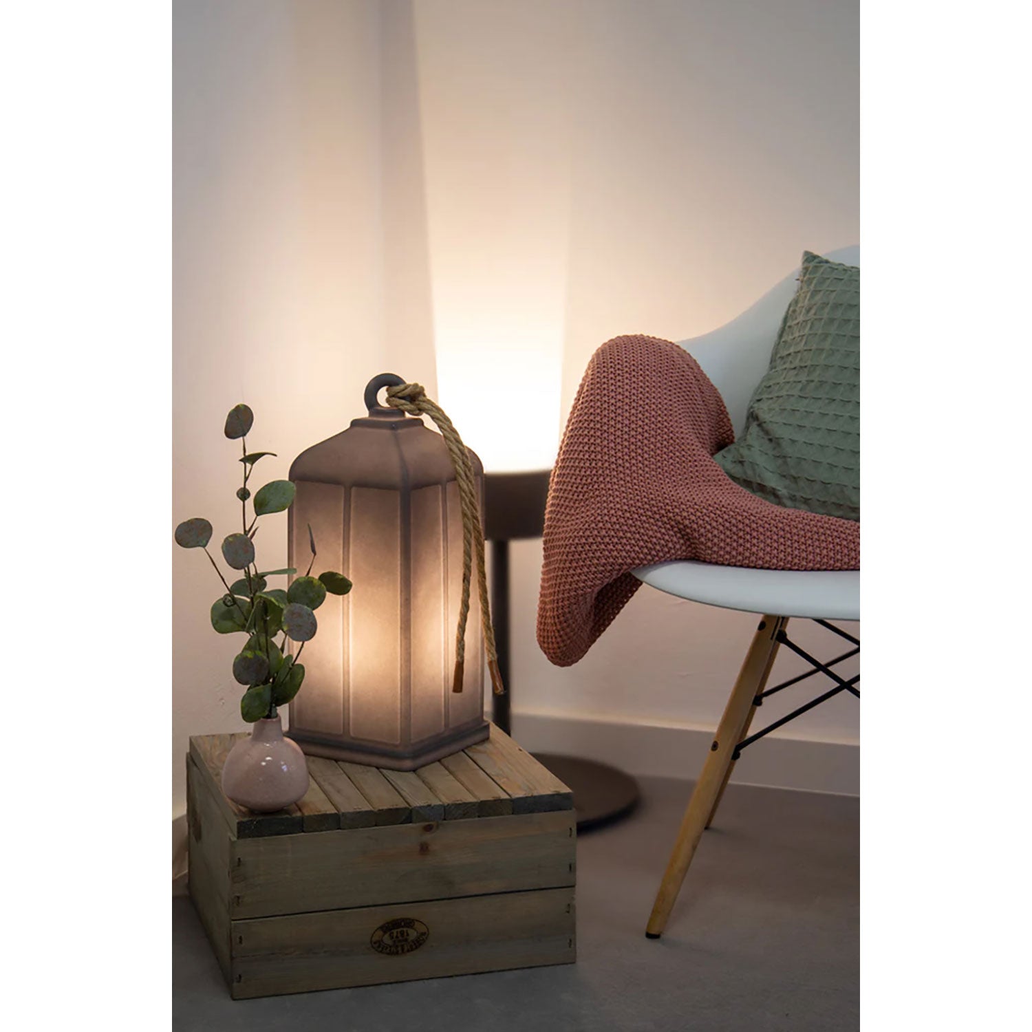 In einer gemütlichen Ecke steht ein weißer Sessel mit einer roten Decke und einem grünen Kissen. Die Leuchtende Solarlaterne leuchtet warm auf einer Holzkiste neben einer kleinen Vase mit Grünpflanzen.