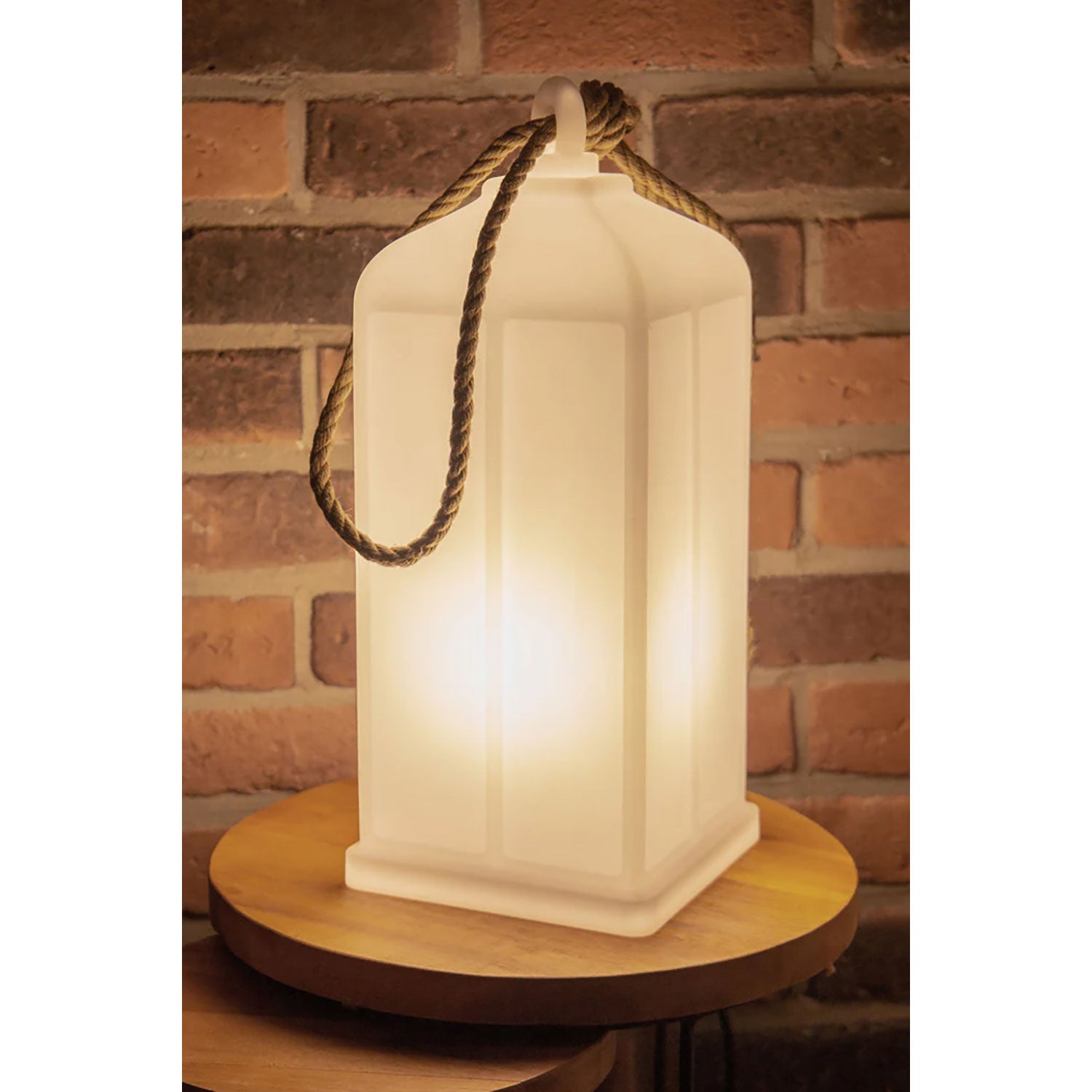 Die leuchtende Solarlaterne mit Seilgriff spendet warmes Licht auf einem runden Holztisch. Ihre rechteckige Form und Wetterbeständigkeit verstärken die gemütliche Atmosphäre vor rustikalen roten Backsteinwänden.