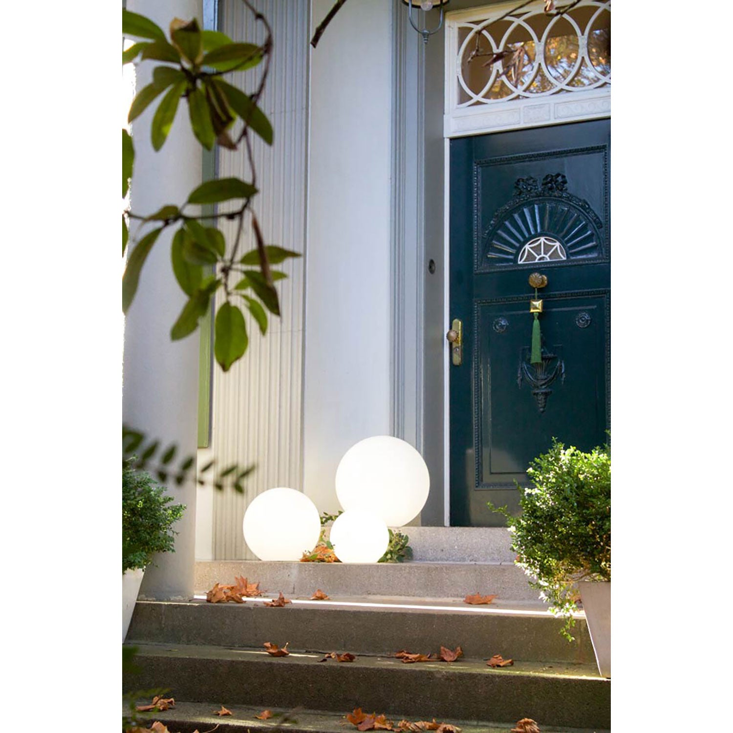 Der stilvolle Eingang besticht durch eine dunkelblaue Tür mit verziertem Glas und Messingknauf. Zwei leuchtende Solar-Leuchtkugeln schmücken die Stufen, umgeben von Laub, flankiert von grünen Büschen in weißen Pflanzgefäßen, während ein belaubter Zweig die linke Seite einrahmt.