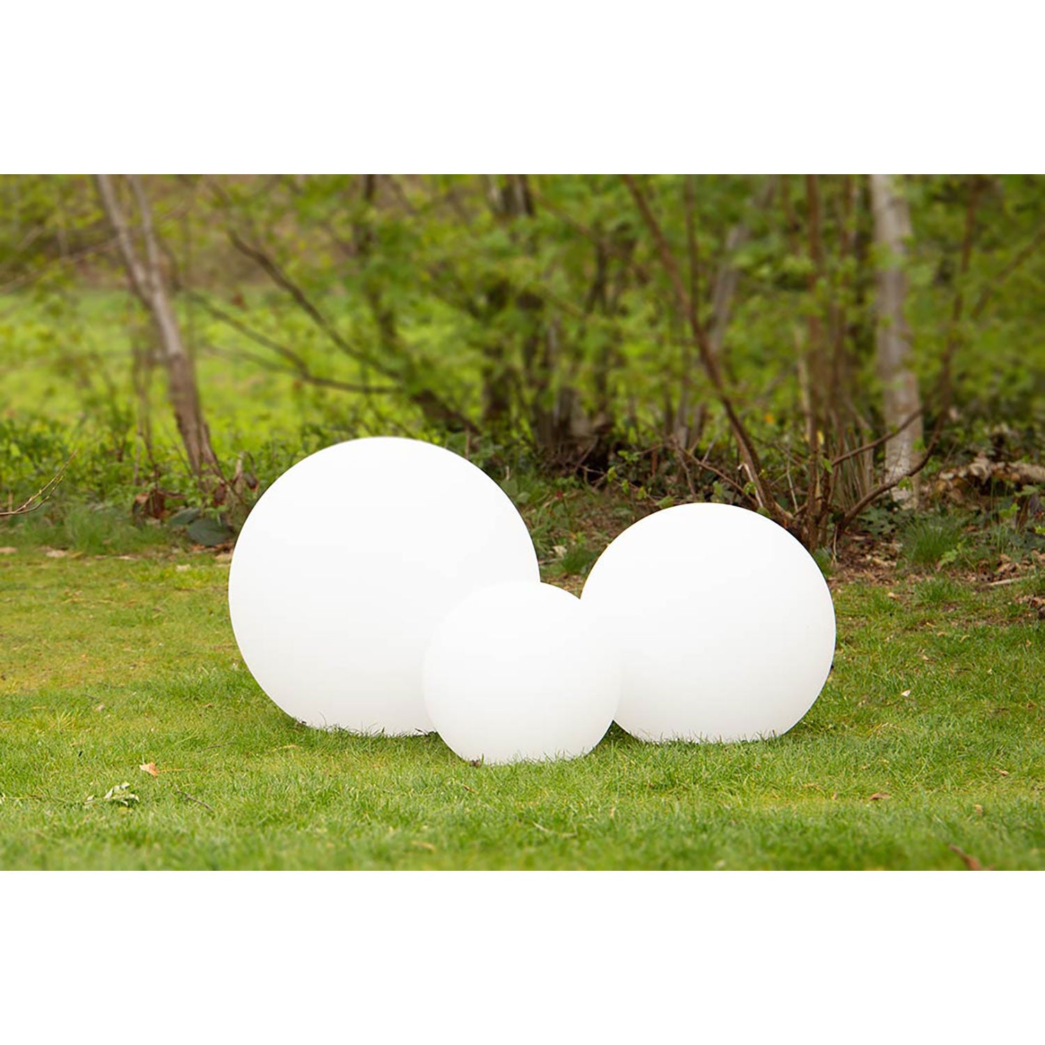 Drei große, leuchtend weiße Leuchtkugeln unterschiedlicher Größe stehen auf einer Wiese mit üppigen grünen Bäumen und Büschen im Hintergrund. Diese eleganten Solarleuchten strahlen ein sanftes Licht aus und schaffen eine ruhige und umweltfreundliche Atmosphäre.