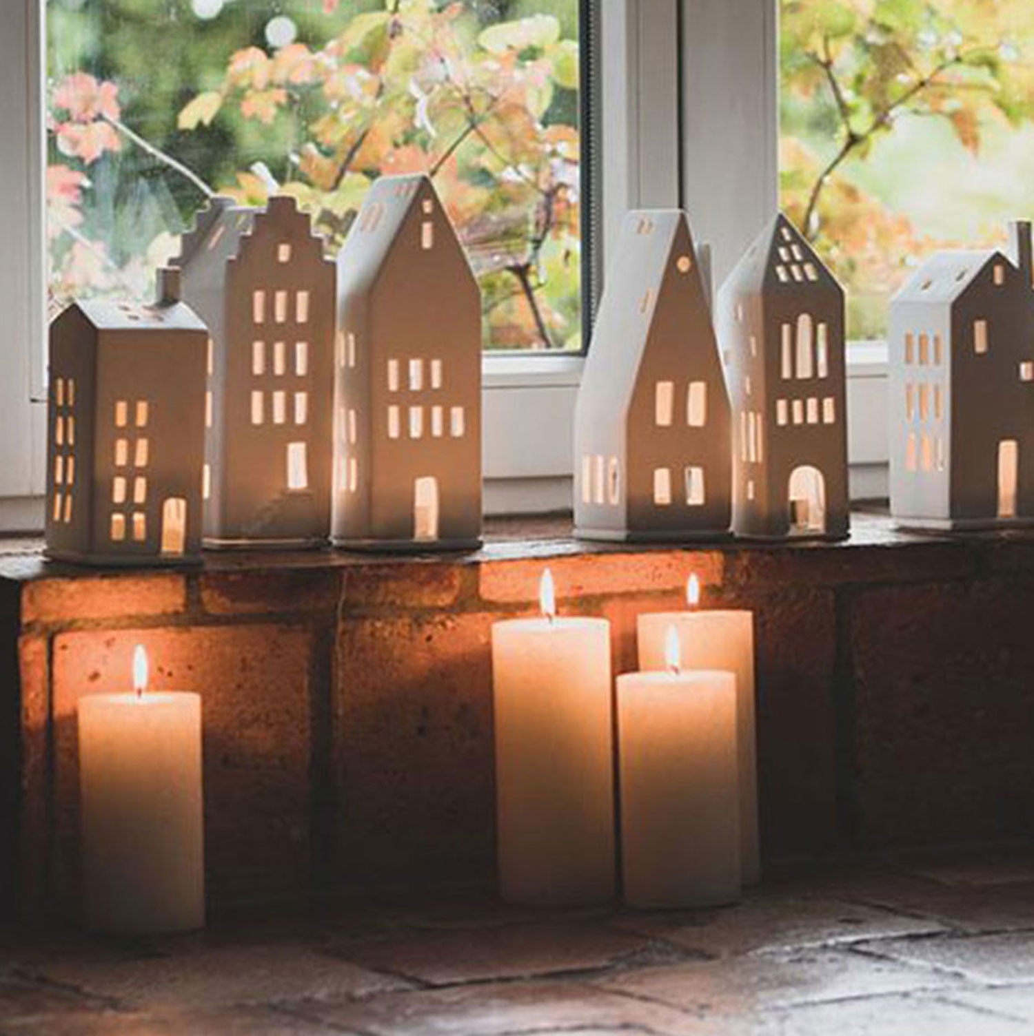 Die Laternen „Mittel, Kleines Fenster“ von Lichthaus strahlen ein warmes Licht auf einer gemauerten Fensterbank aus. Drei brennende Kerzen unterschiedlicher Höhe stehen davor. Handarbeit verstärkt den Charme, und das verschwommene Grün rundet die ideale Weihnachtsdekoration ab.