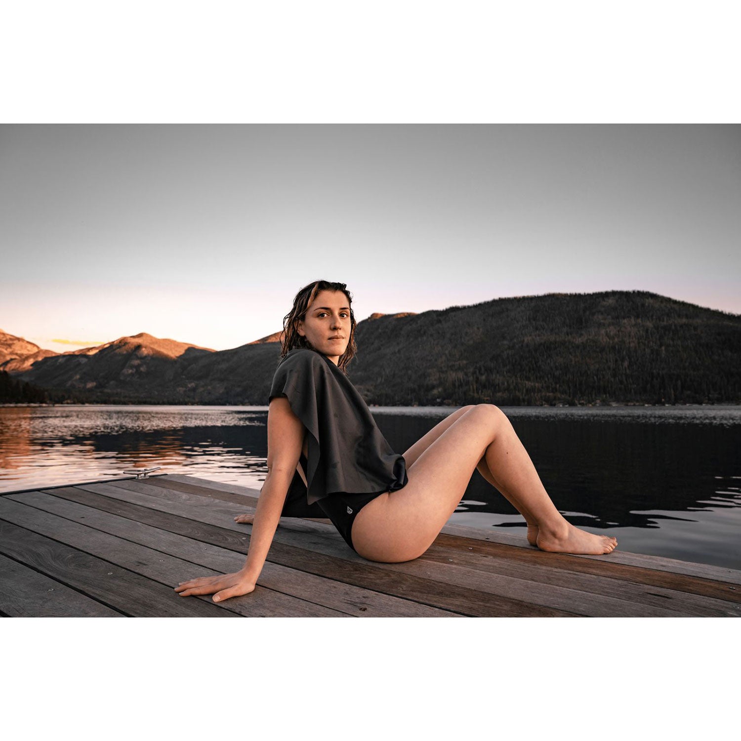 Eine Person sitzt bei Sonnenuntergang auf einem Holzsteg am See, eingehüllt in ein ultraleichtes Nanodry Handtuch über ihrer Badebekleidung. Die Berge spiegeln sich im ruhigen Wasser unter klarem Himmel, während die antimikrobielle Beschichtung des Handtuchs für Frische und Komfort sorgt.