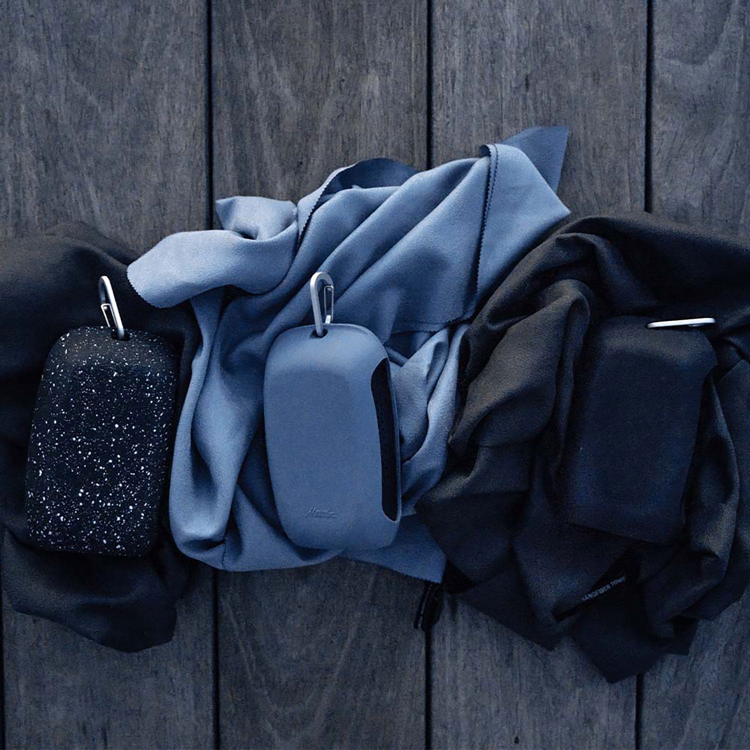 Drei tragbare Lautsprecher des Ultraleichtes Nanodry Handtuchs, jeweils in einer eigenen Silikonhülle mit antimikrobieller Beschichtung, liegen auf blauem und schwarzem, zerknittertem Stoff. Ihre Karabinerhaken sind sichtbar.