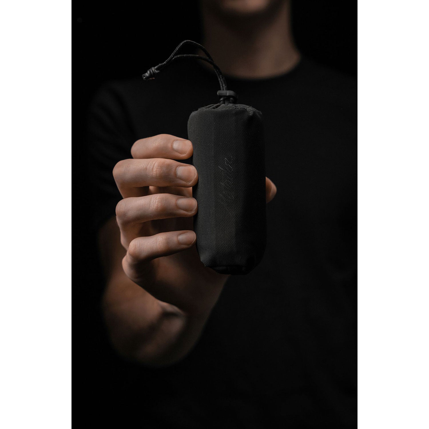 Eine Person in einem schwarzen Hemd hält den schlanken, zylindrischen Beutel des ultraleichten Nanodry Handtuchs mit Kordelzug vor einem schattigen Hintergrund. Die antimikrobielle Beschichtung hält den Beutel frisch und sauber – ideal zum Schutz Ihrer wichtigsten Dinge. Das Gesicht der Person ist durch die schwache Beleuchtung verdeckt.