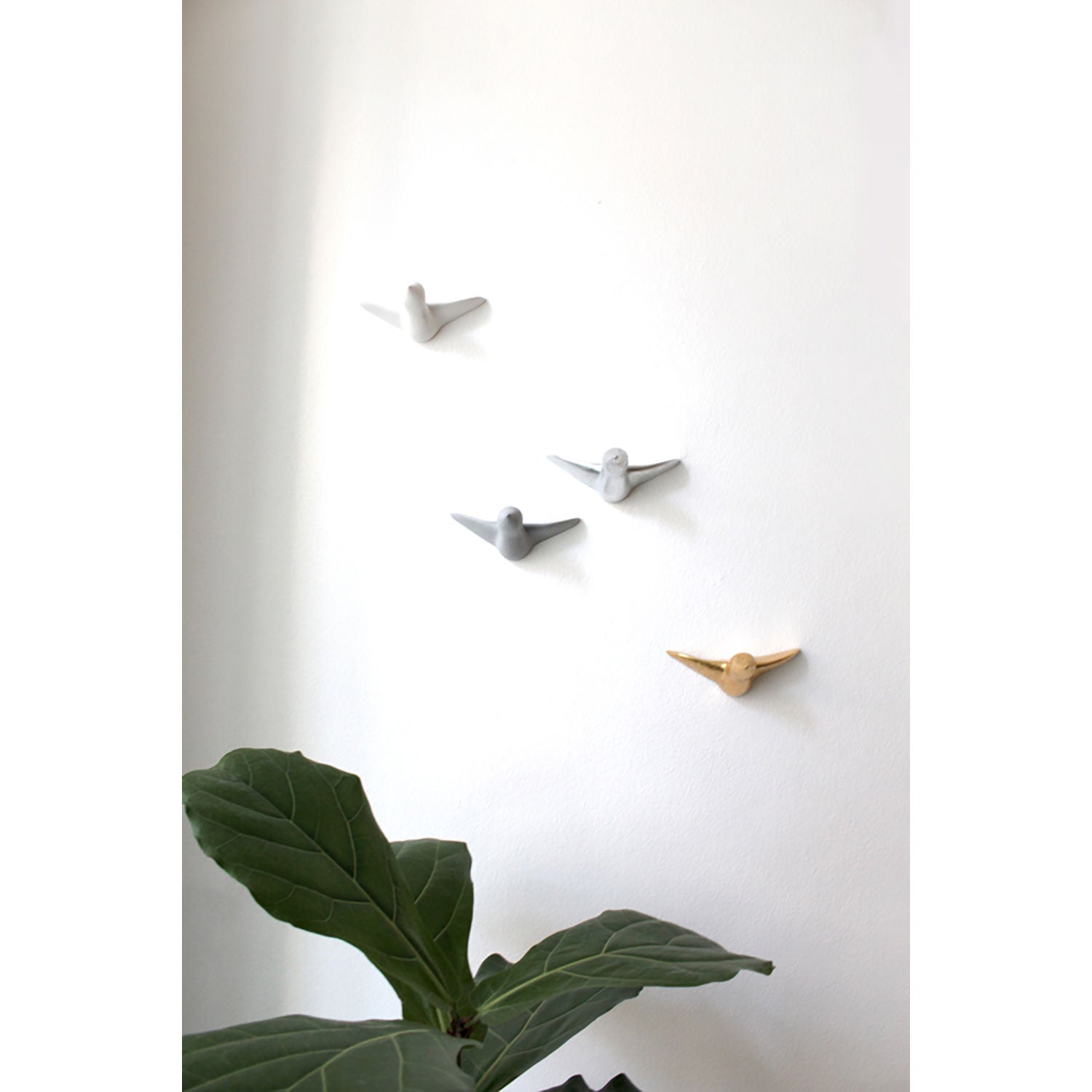 Vier vogelförmige Wandhaken (Wandskulptur - gemischter Vogelschwarm) in Weiß, Gold, Grau und Silber zieren eine weiße Wand. In der unteren linken Ecke schmiegt sich eine üppige grüne Pflanze mit großen Blättern an und verleiht jedem Raum mit ihrem eleganten Design und ihrer Handwerkskunst Charme.