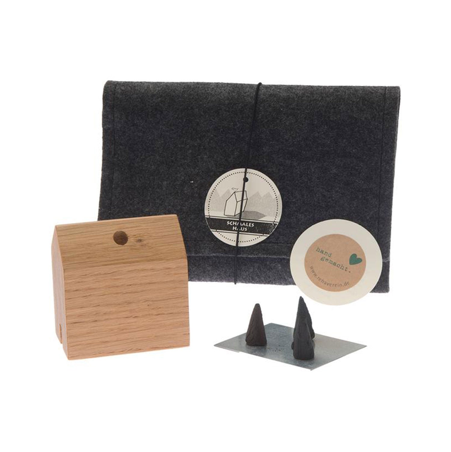 Das „Räucherhäuschen Schmales Haus“ ist ein Holzblock mit Loch in einem dunkelgrauen Lederetui mit Hülle. Darin befinden sich runde weiße Etiketten und zwei kleine kegelförmige Objekte auf einem grauen rechteckigen Ständer, die perfekt dazu geeignet sind, den für die Weihnachtstradition typischen, gemütlichen Duft von Tannenduftkegeln hervorzurufen.