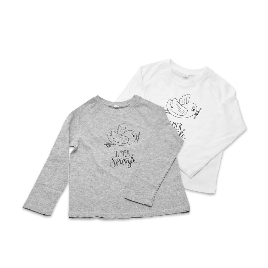 Zwei langärmelige Kindershirts, grau und weiß, beide mit handgeschriebenem "Ulmer Spätzle"-Schriftzug und einer Vogelillustration auf der Vorderseite. Produkt: Shirt "Ulmer Spätzle", 4-5 Jahre.